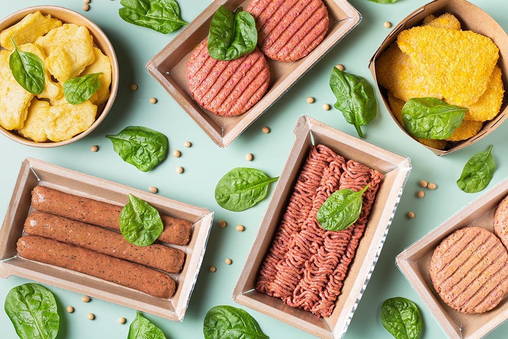 Assortment of plant-based meat alternatives in cardboard trays, including burger patties, sausages, ground meat, nuggets, and breaded cutlets, arranged on a light green background with spinach leaves and soybea