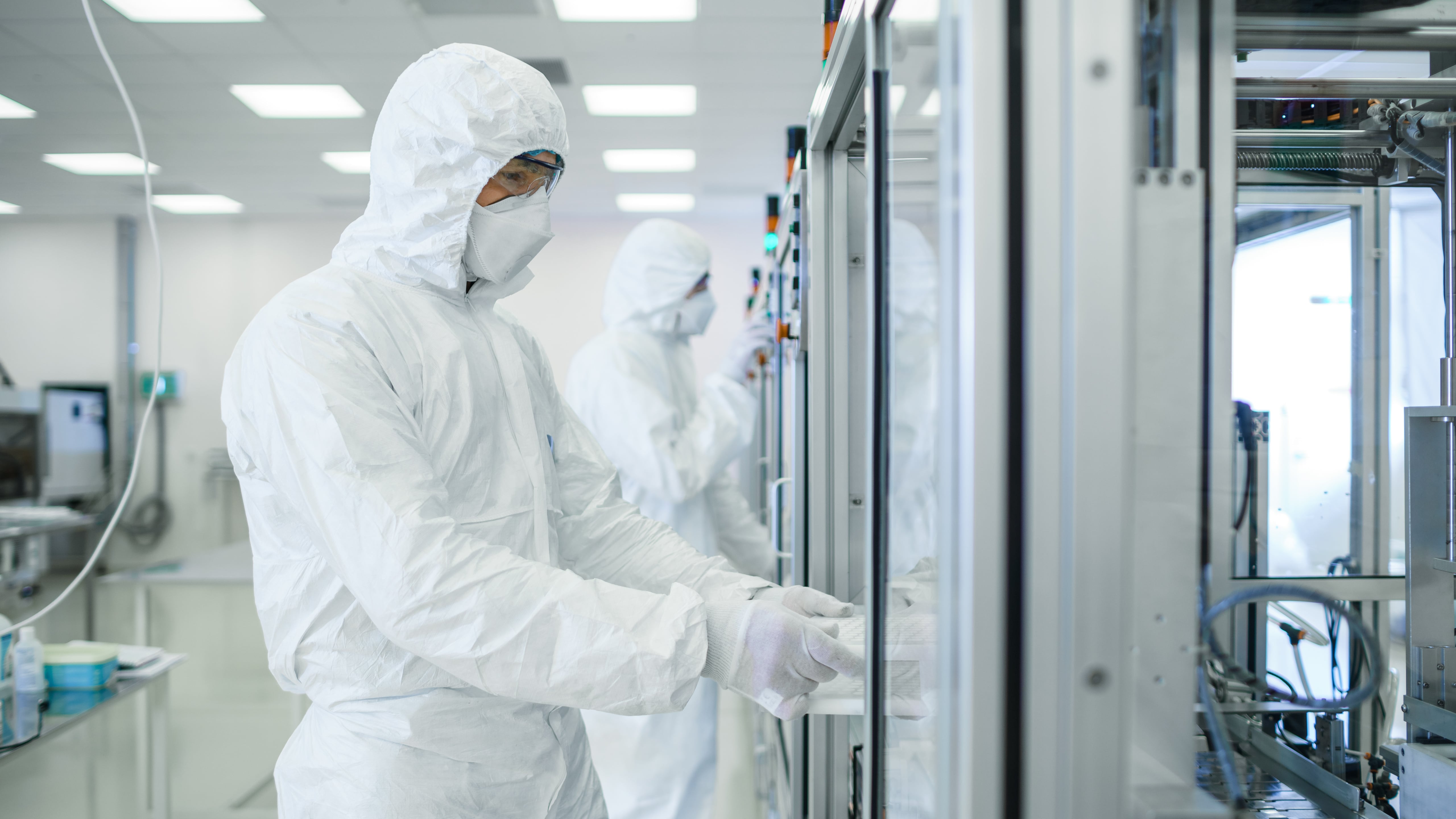 Two individuals wearing full-body cleanroom suits and face coverings working inside a sterile laboratory environment, operating equipment with glass panels and control buttons.