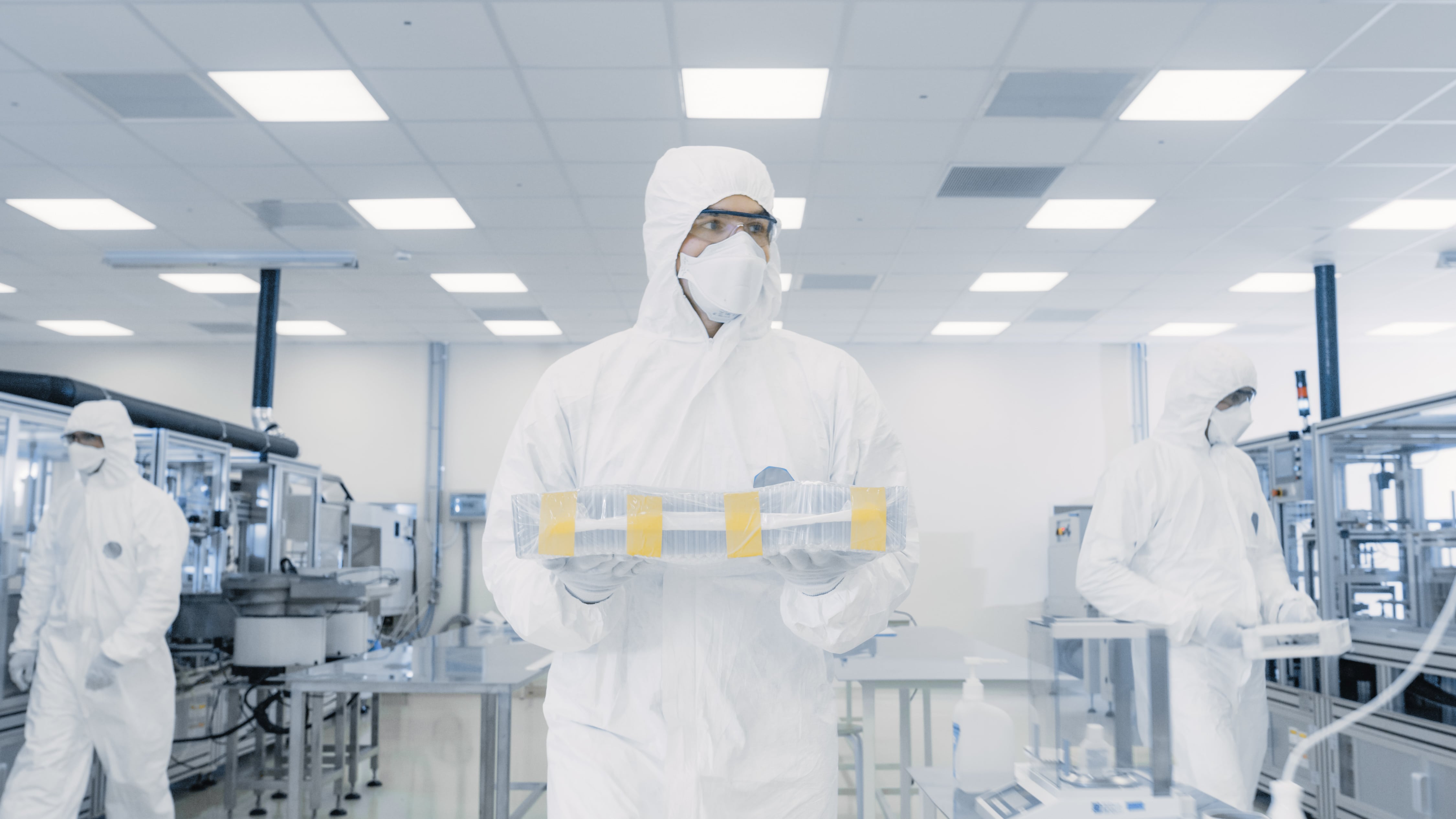 Three individuals wearing full-body cleanroom suits and face masks inside a bright, sterile laboratory. The person in the center is holding a tray with multiple sealed containers, while others work near advanced machinery and equipment in the background.