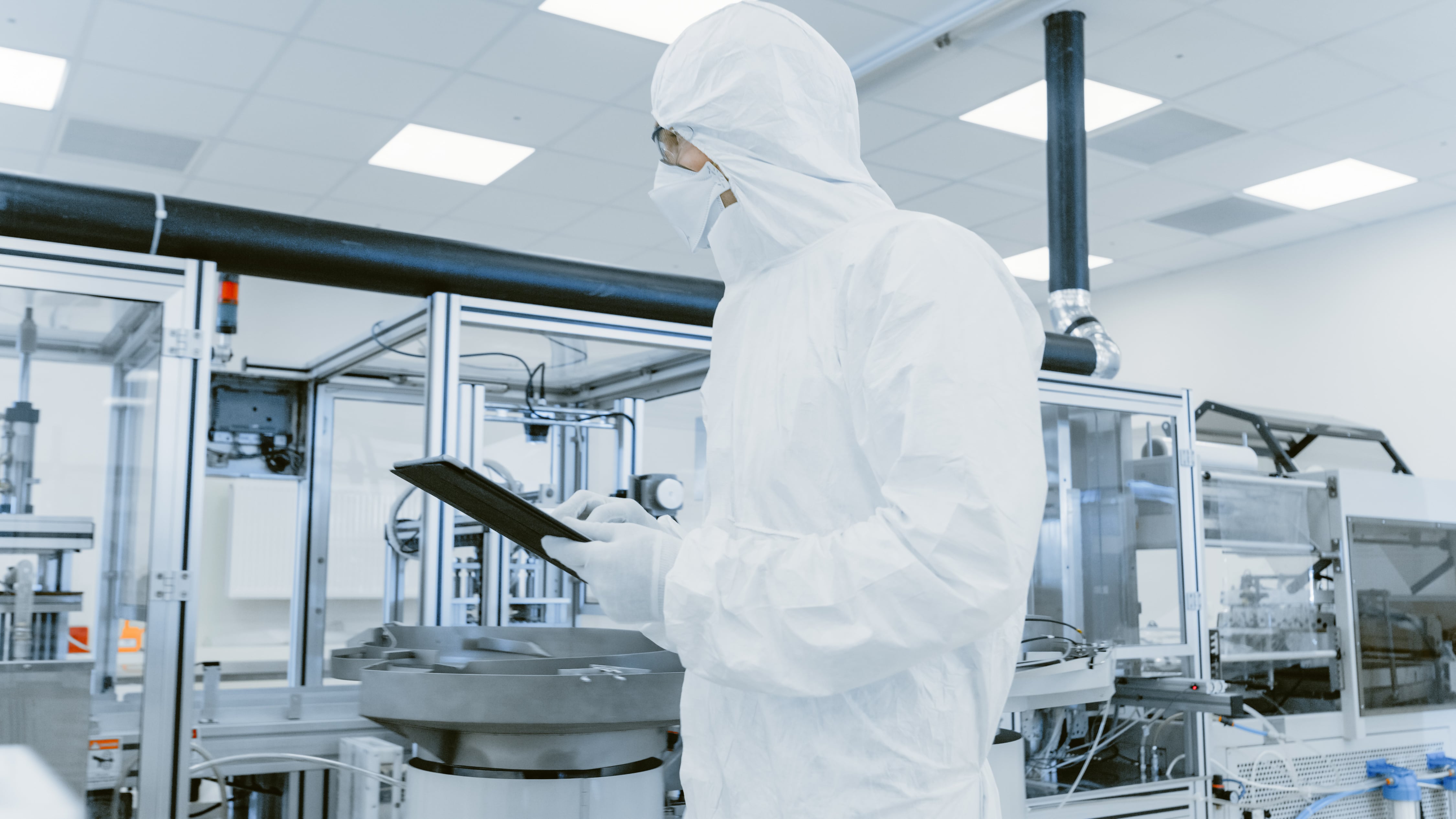 Person wearing a full-body cleanroom suit and face mask inside a sterile laboratory, holding a tablet while inspecting advanced machinery with glass panels and metal components.