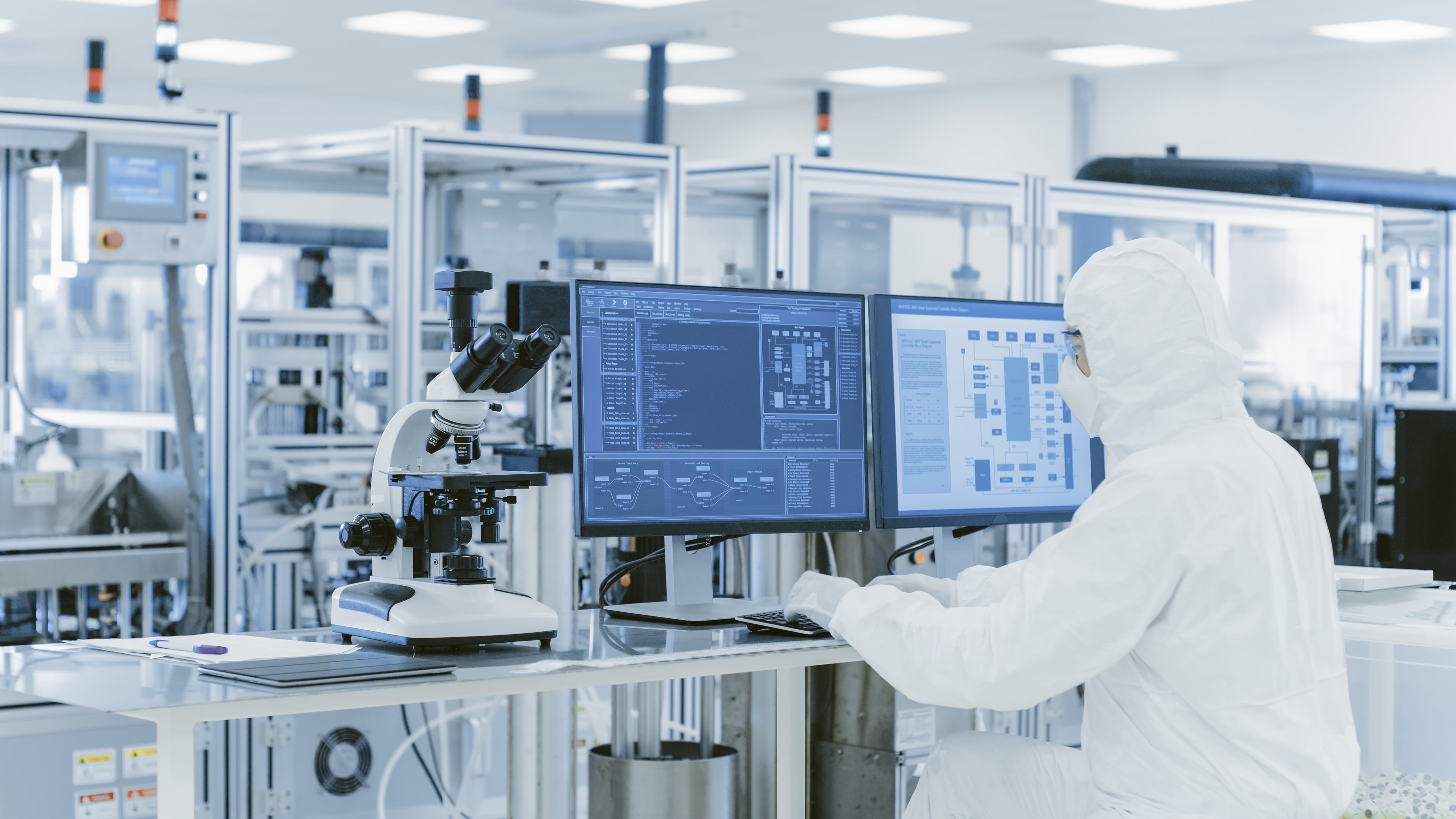 Person in a full-body cleanroom suit seated at a workstation inside a sterile laboratory, operating two large computer monitors displaying technical diagrams and data, with a microscope and advanced machinery in the background.