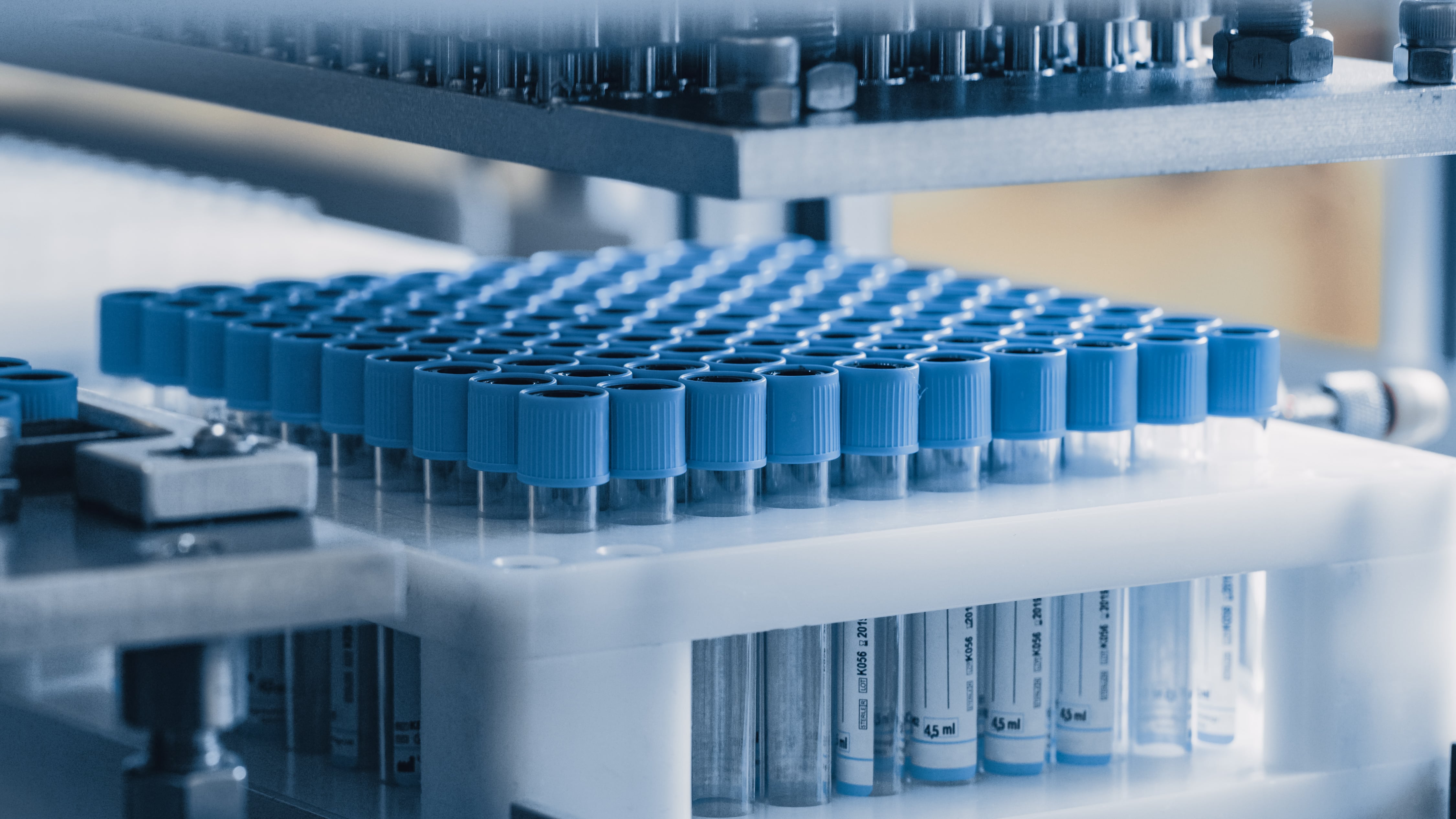 Close-up of automated laboratory equipment holding a tray filled with multiple test tubes sealed with blue caps, arranged in neat rows, inside a sterile production environment.