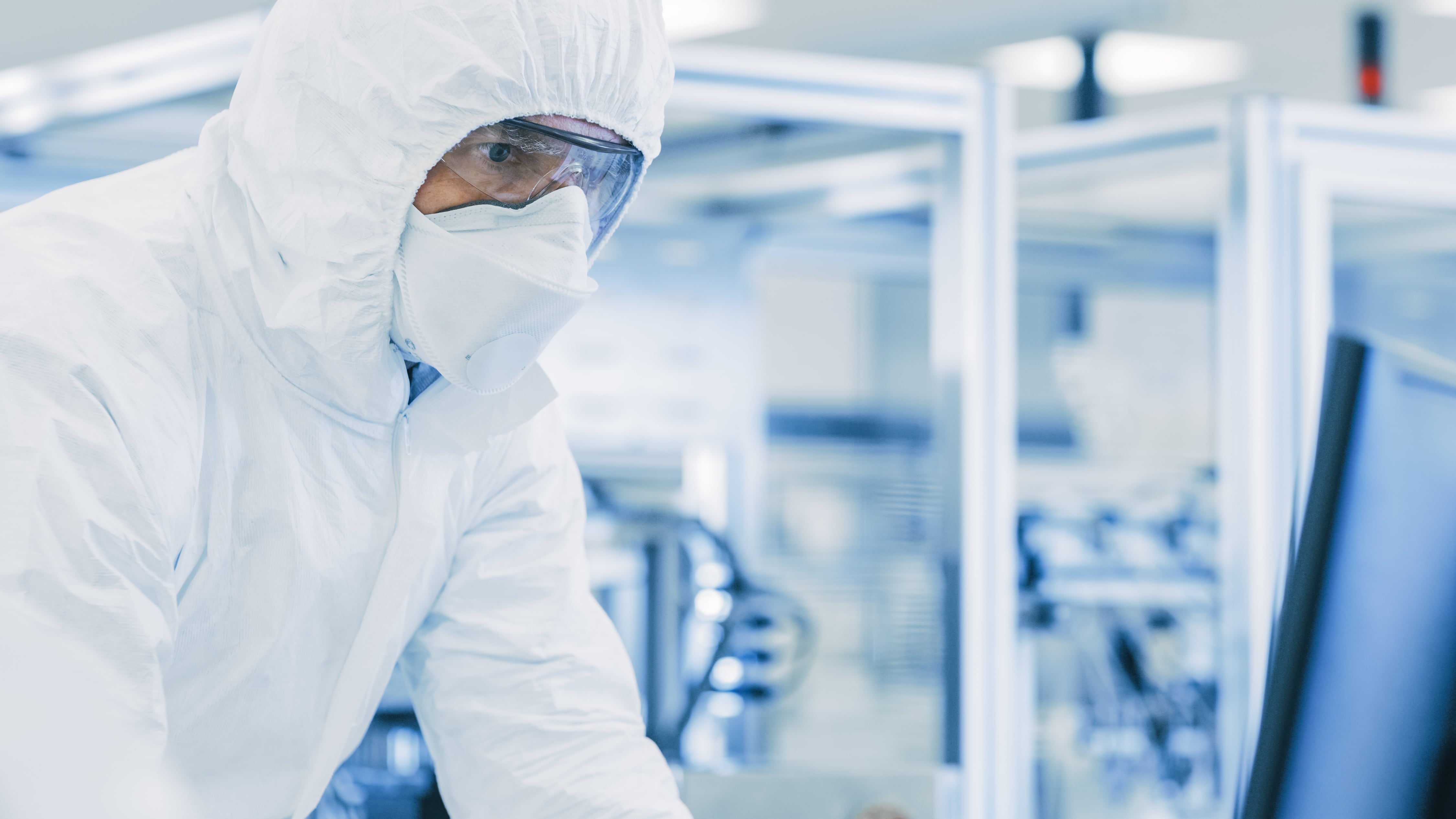 Close-up of a person wearing a full-body cleanroom suit and protective goggles inside a sterile laboratory, working near advanced equipment and computer monitors.