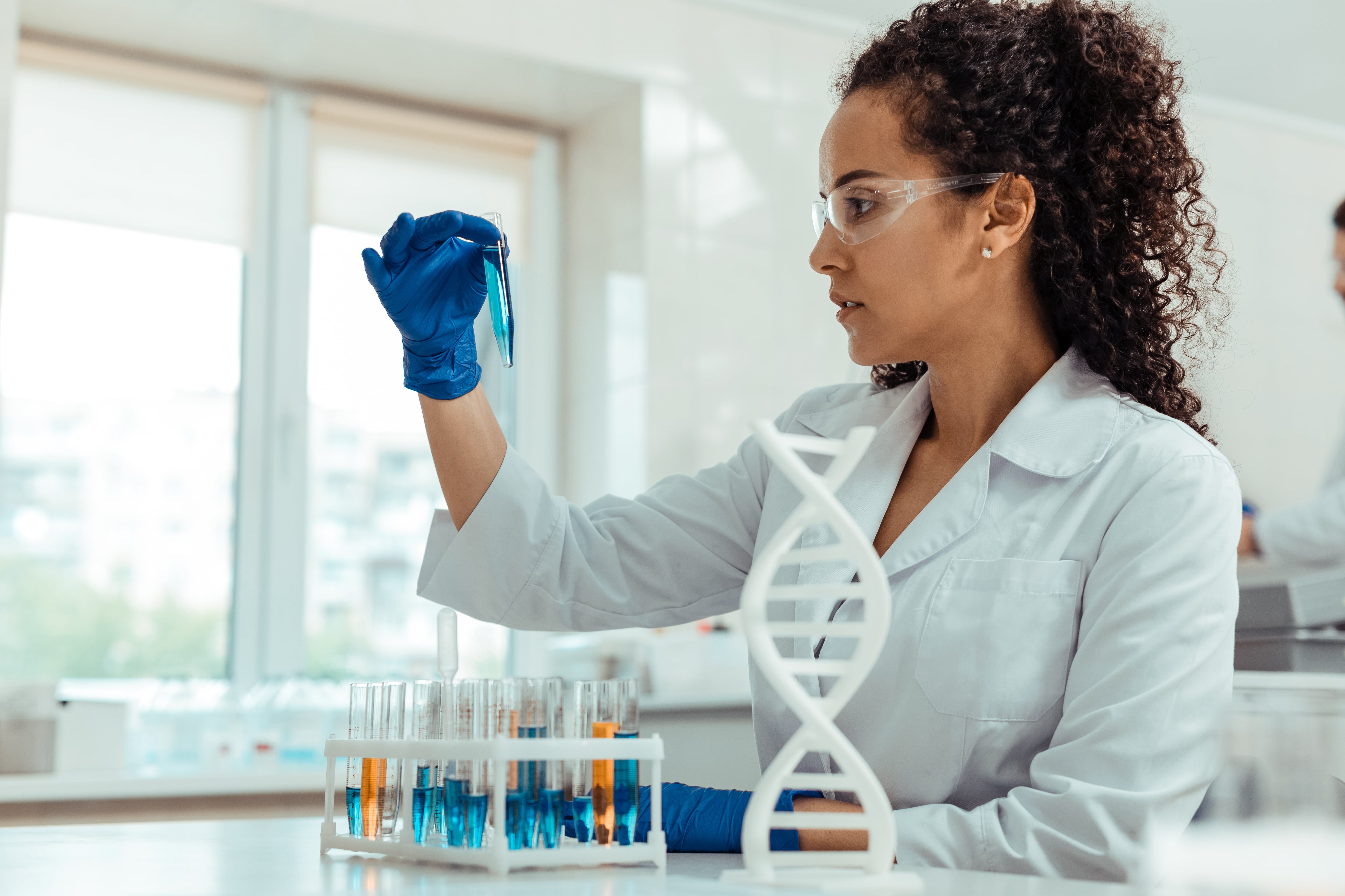 Scientist wearing a white lab coat and blue gloves holding a test tube with blue liquid in a laboratory setting, with several other test tubes and a DNA model on the table.