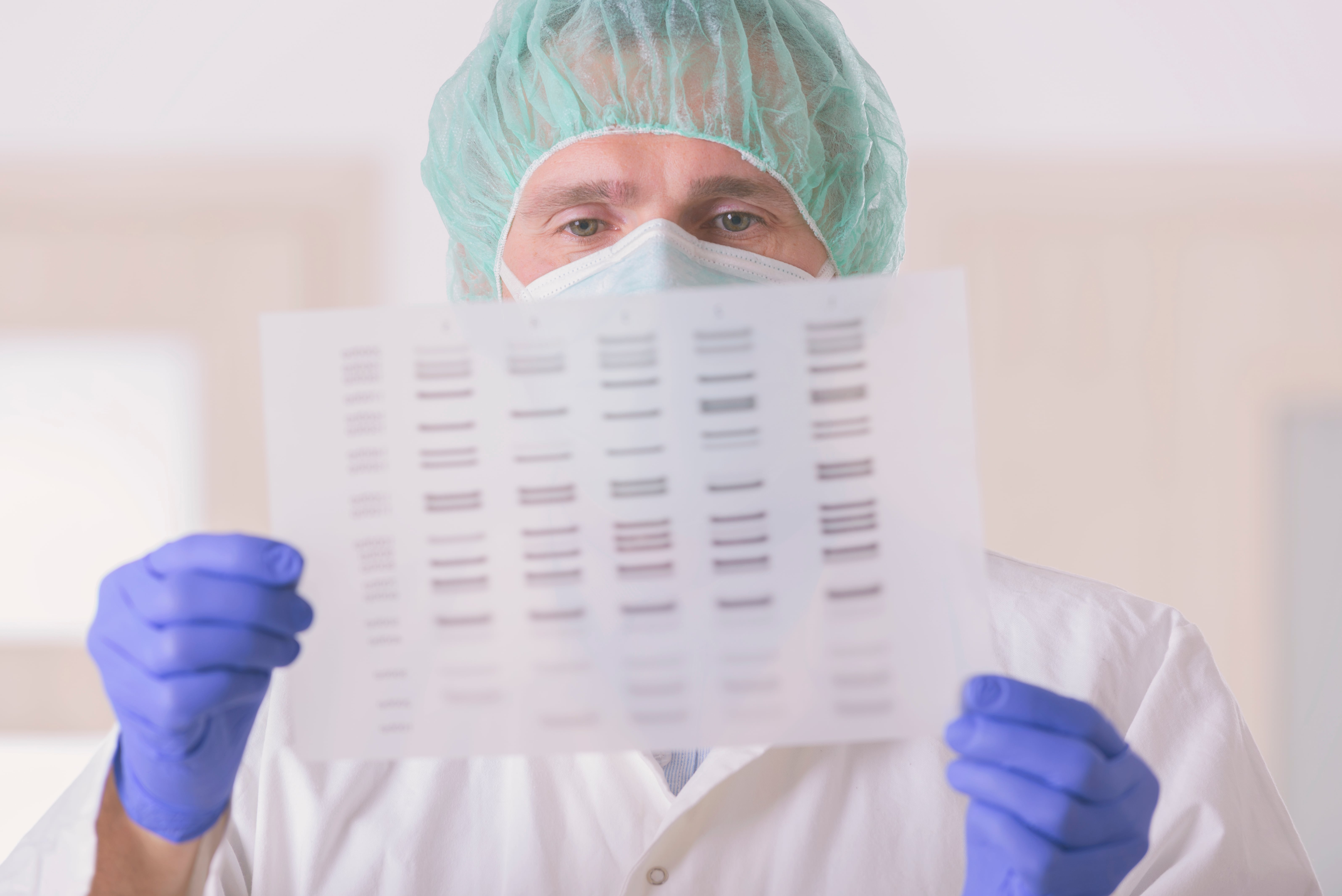 Laboratory professional wearing protective gloves and a hair cover holding a transparent sheet with printed gel electrophoresis bands in a scientific setting.