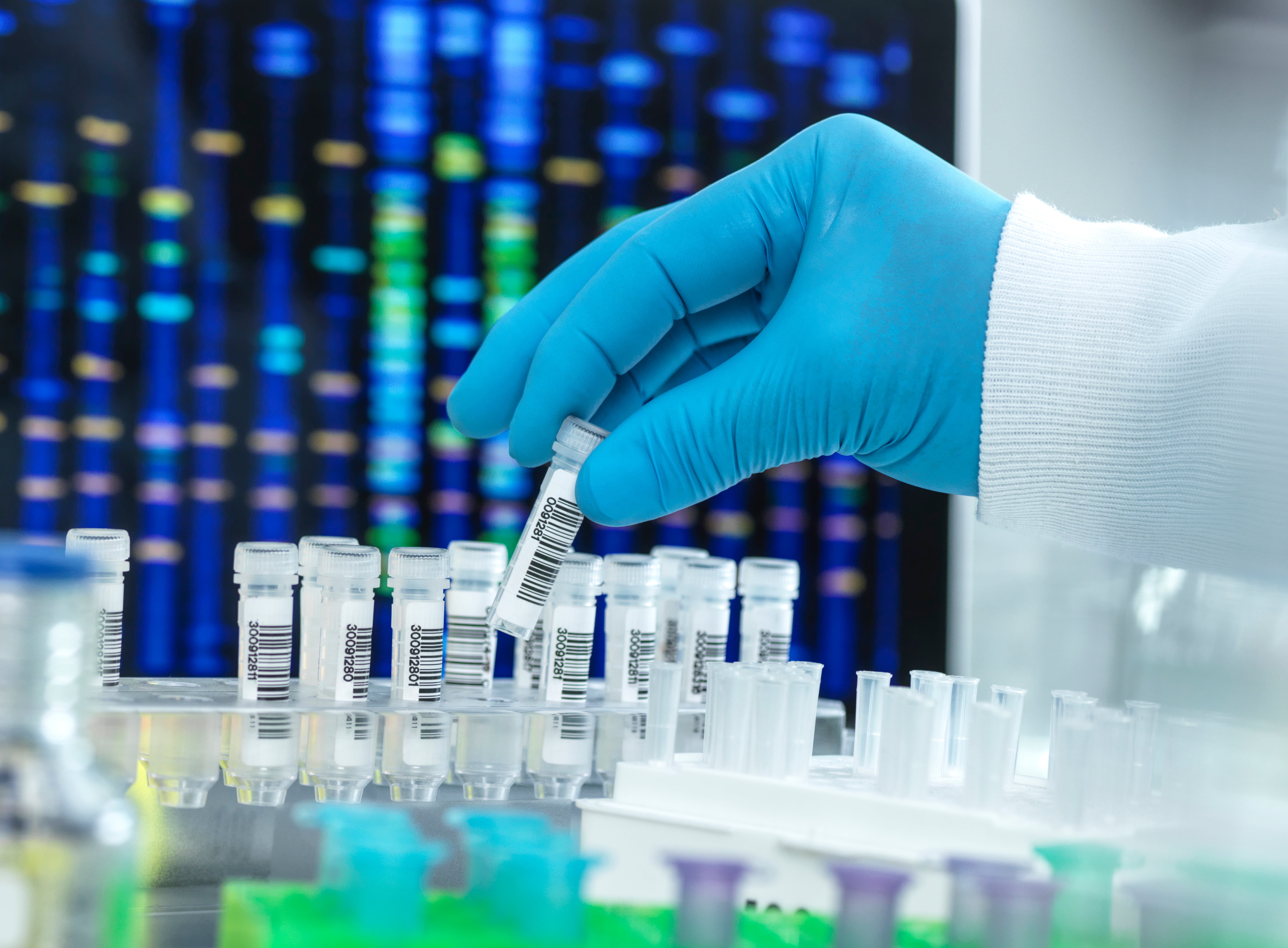 Gloved hand placing a small test tube into a rack inside a laboratory, with colorful DNA sequencing patterns displayed on a monitor in the background.