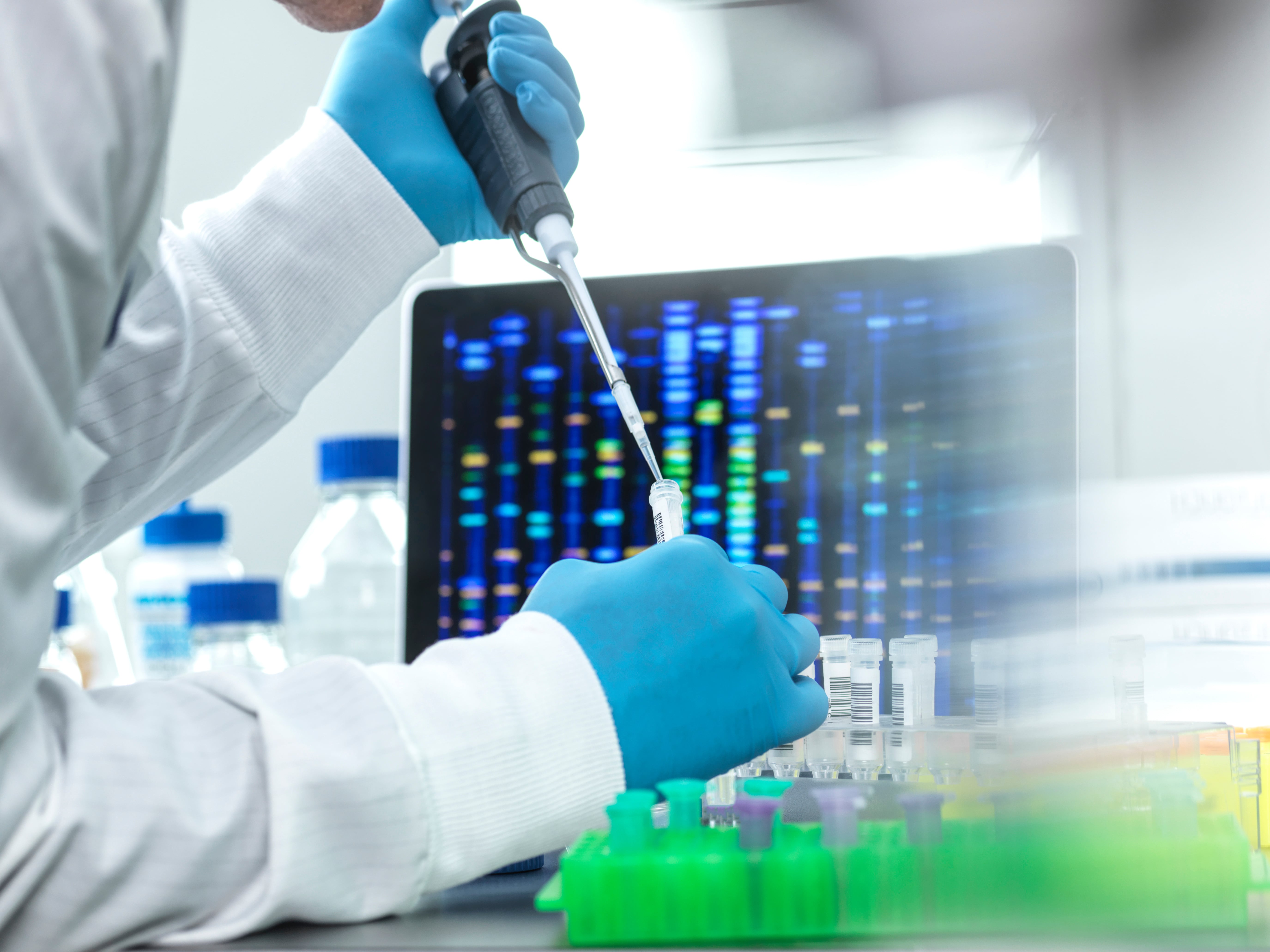 Laboratory technician wearing blue gloves using a pipette to transfer liquid samples into small tubes, with a computer screen in the background displaying colorful DNA sequencing patterns.