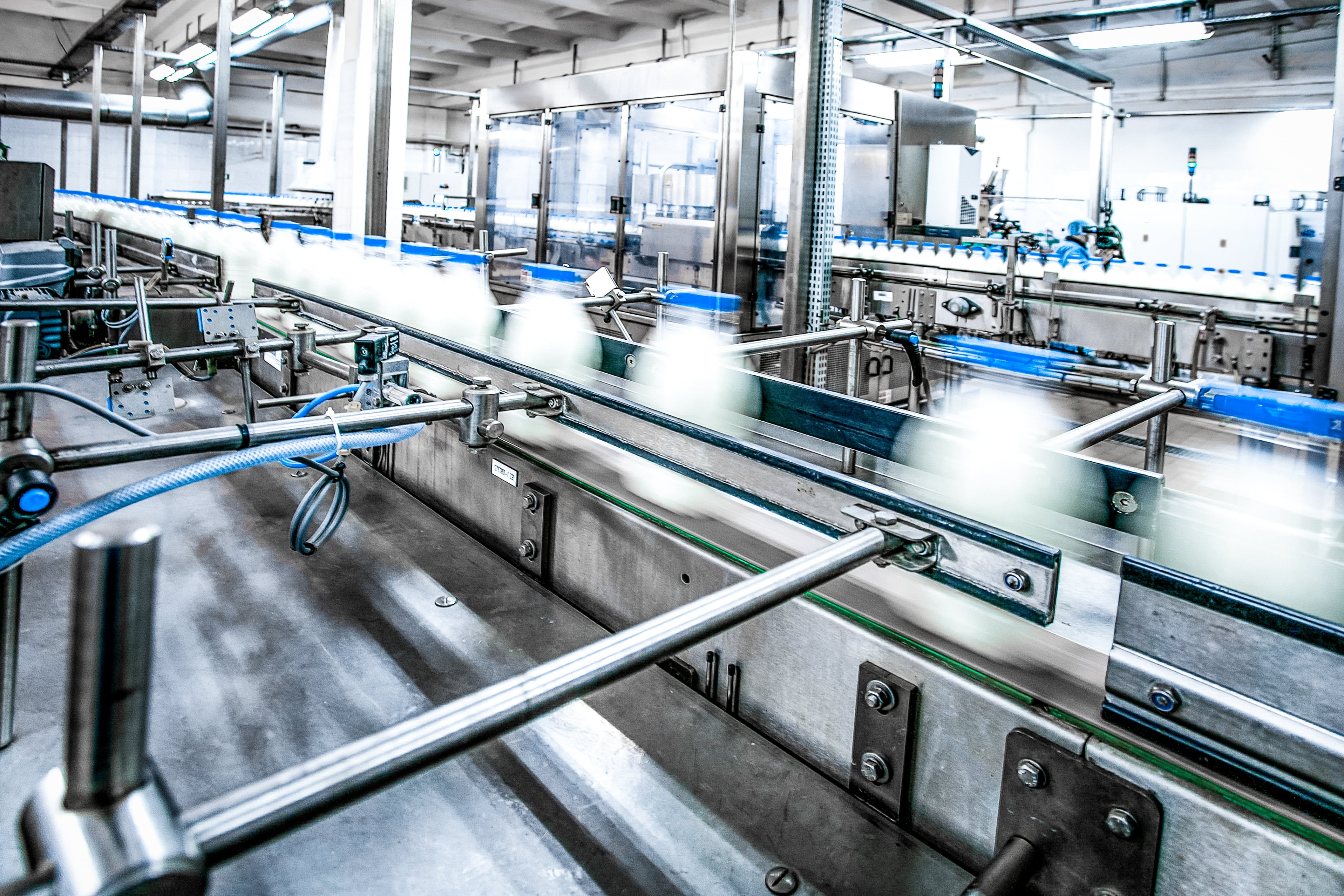Industrial production line with white bottles moving quickly on a conveyor belt inside a modern factory, surrounded by stainless steel machinery and automated equipment.