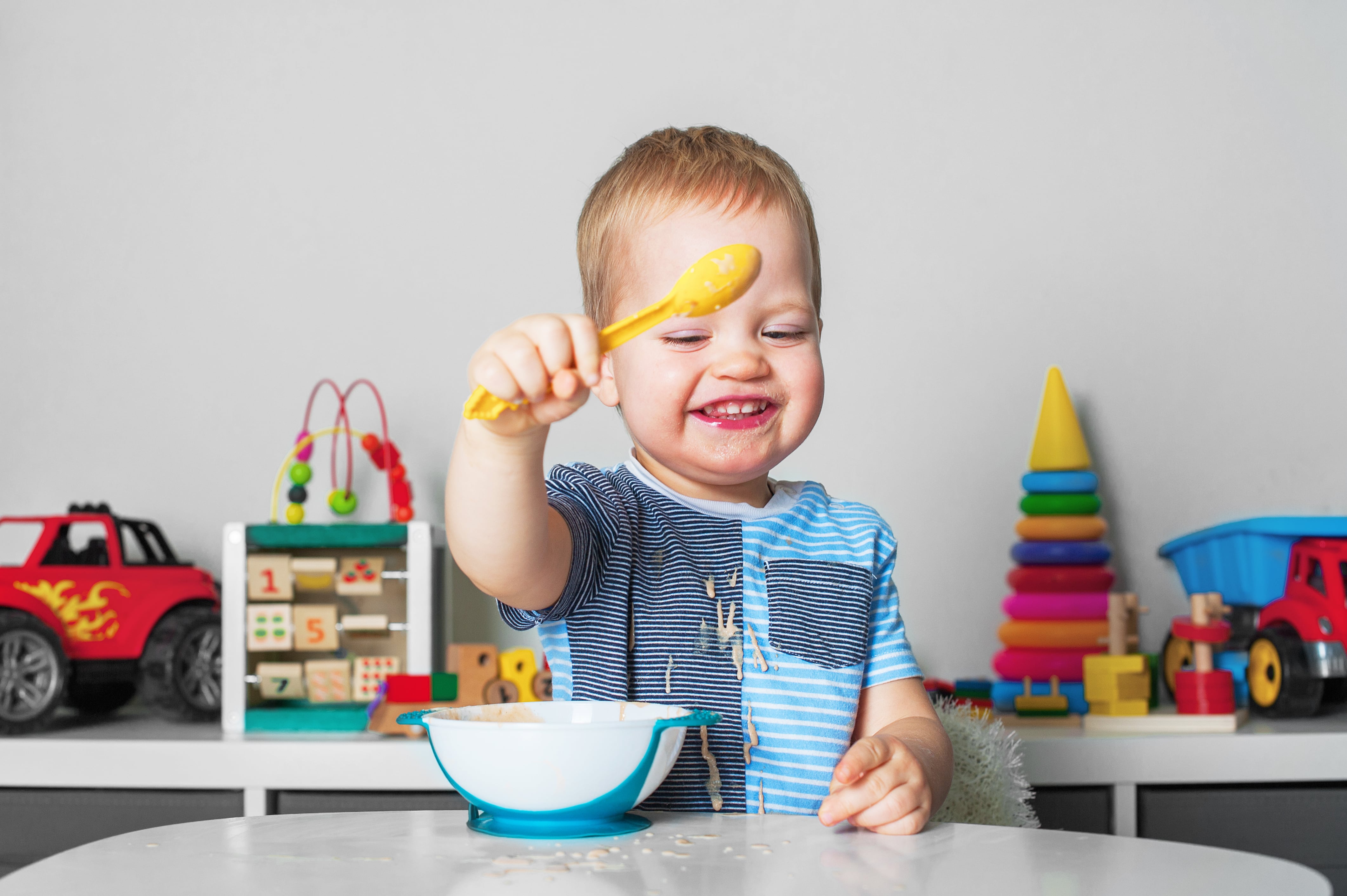Child sitting at a table holding a piece of food above a bowl, with colorful toys such as cars and stacked blocks in the background.