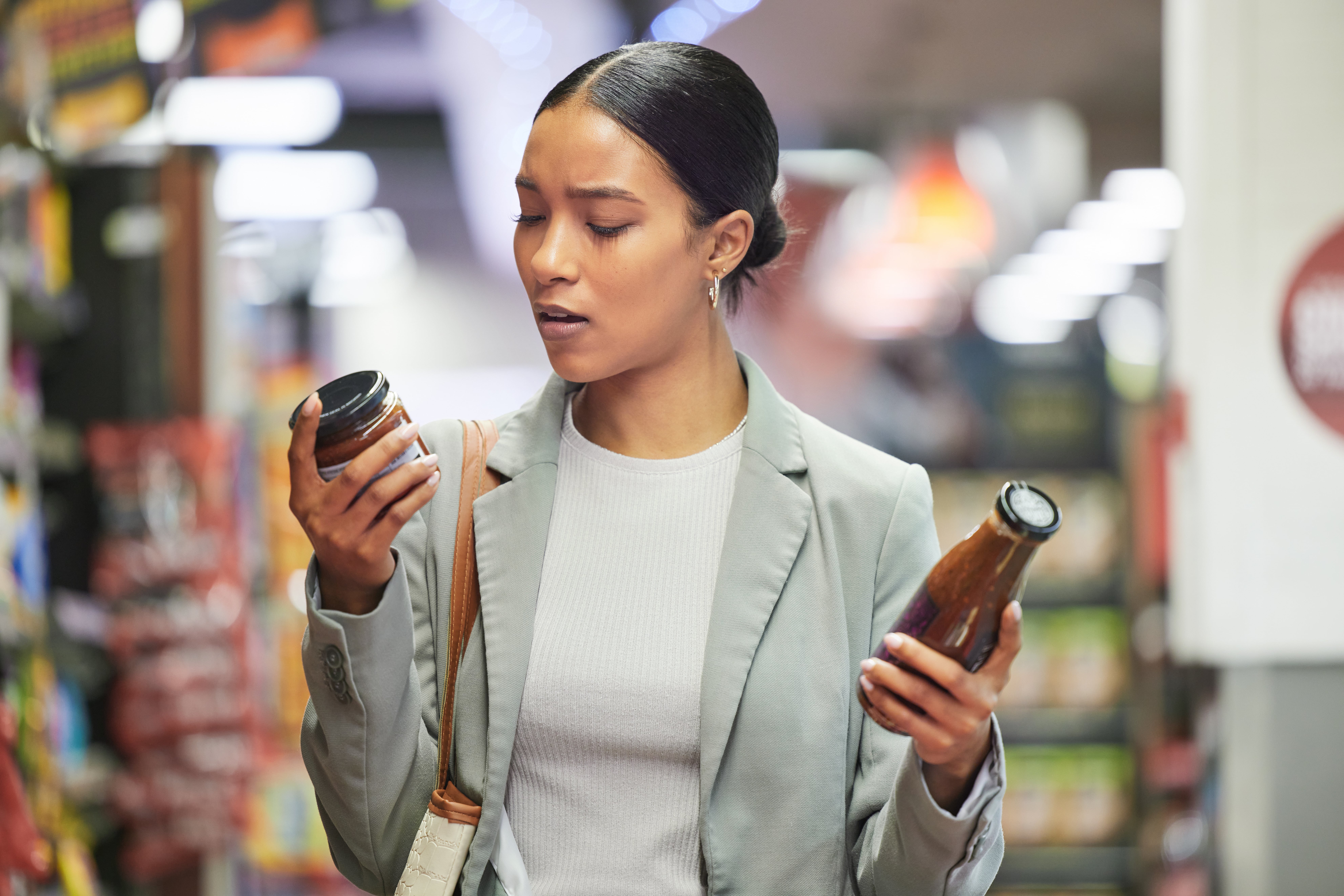 Person in a supermarket holding two bottles, comparing products in an aisle with shelves stocked with items.