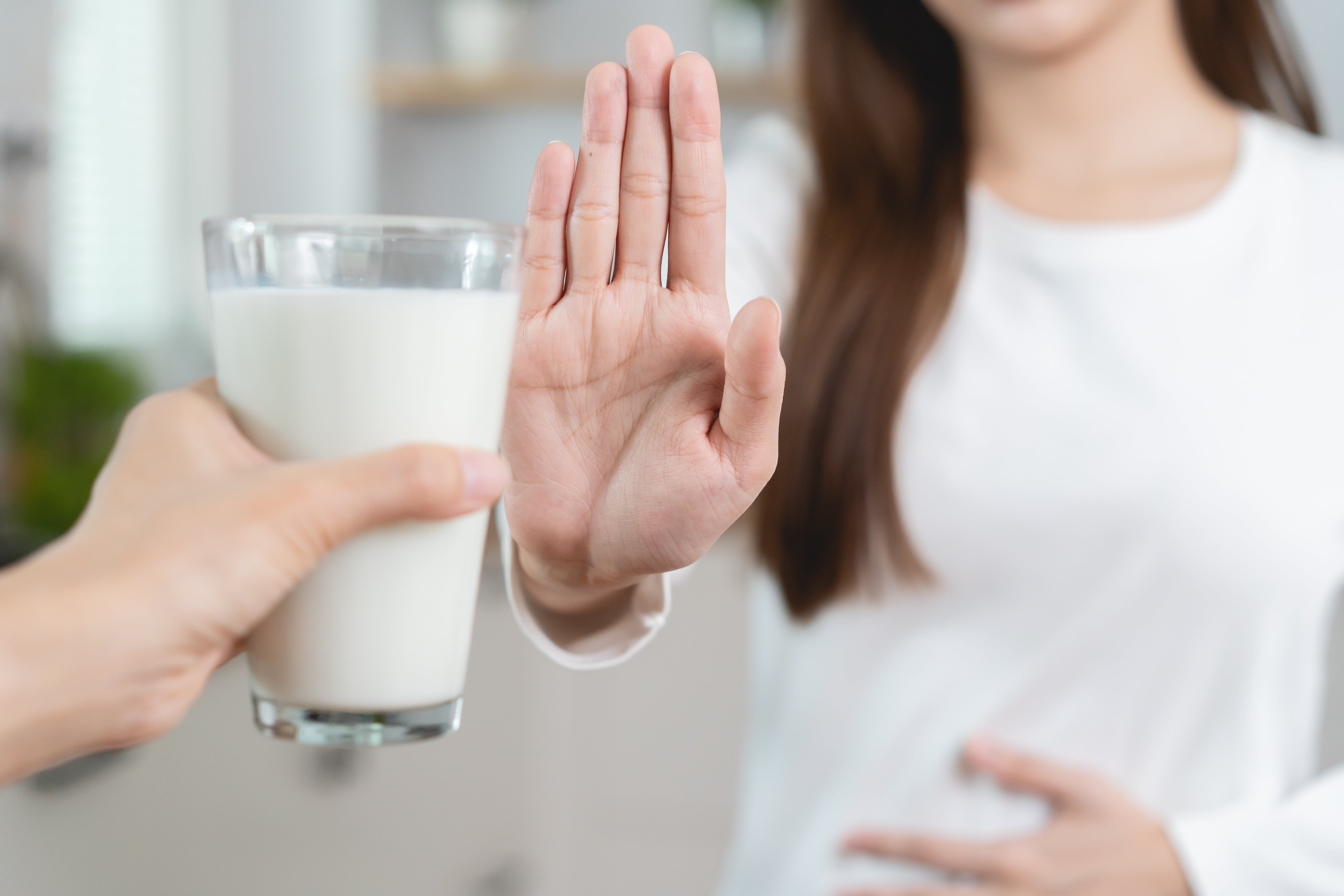 Person refusing a glass of milk with an outstretched hand while placing the other hand on the abdomen, in a home setting.