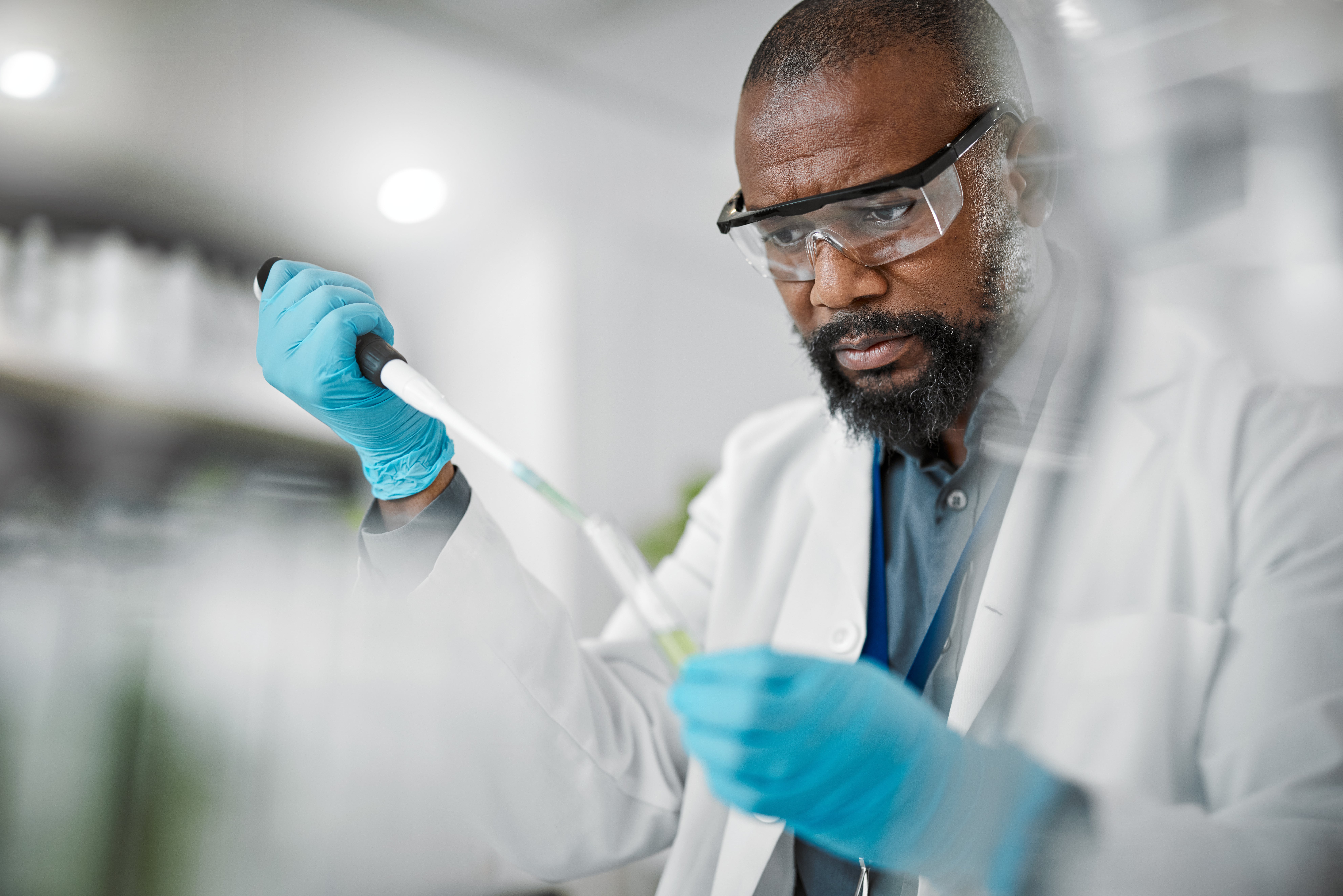 Scientist wearing a white lab coat and blue gloves using a pipette in a laboratory setting, with blurred laboratory equipment in the background.