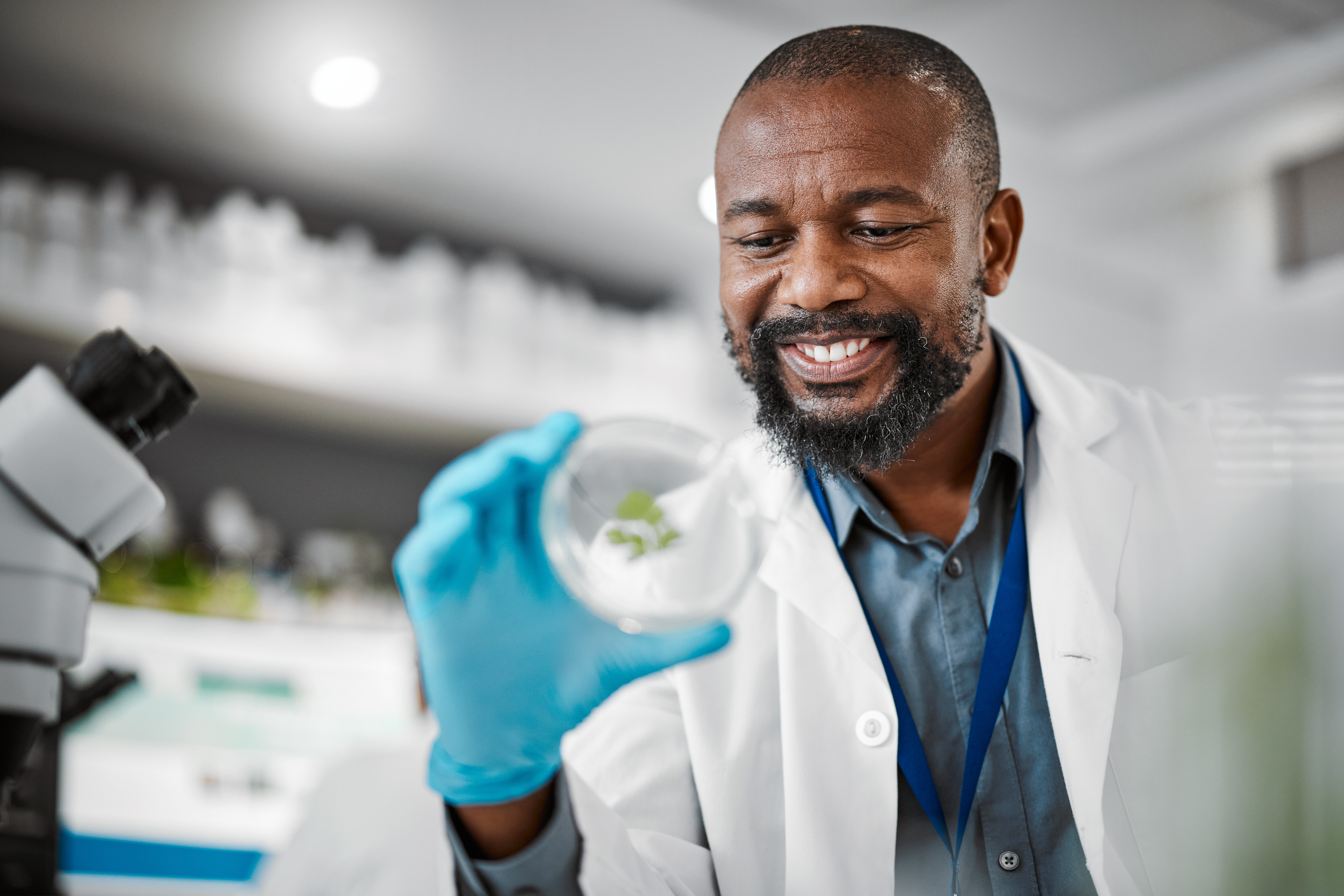 Scientist in a laboratory holding a Petri dish with blue gloves, next to a microscope, with shelves of lab equipment in the background.