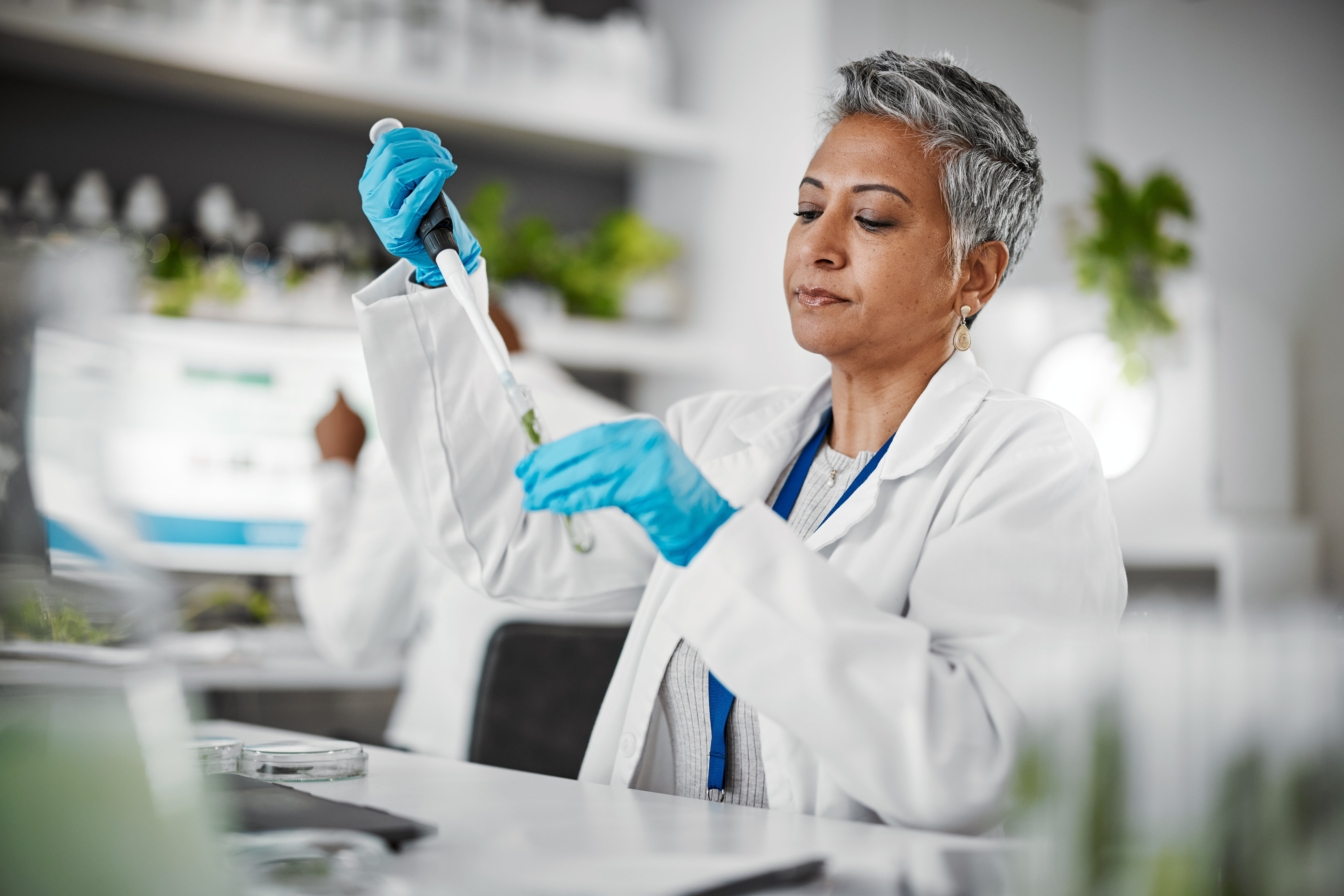 Scientist in a laboratory wearing a white coat and blue gloves, carefully using a pipette to transfer liquid, with shelves of lab equipment in the background.