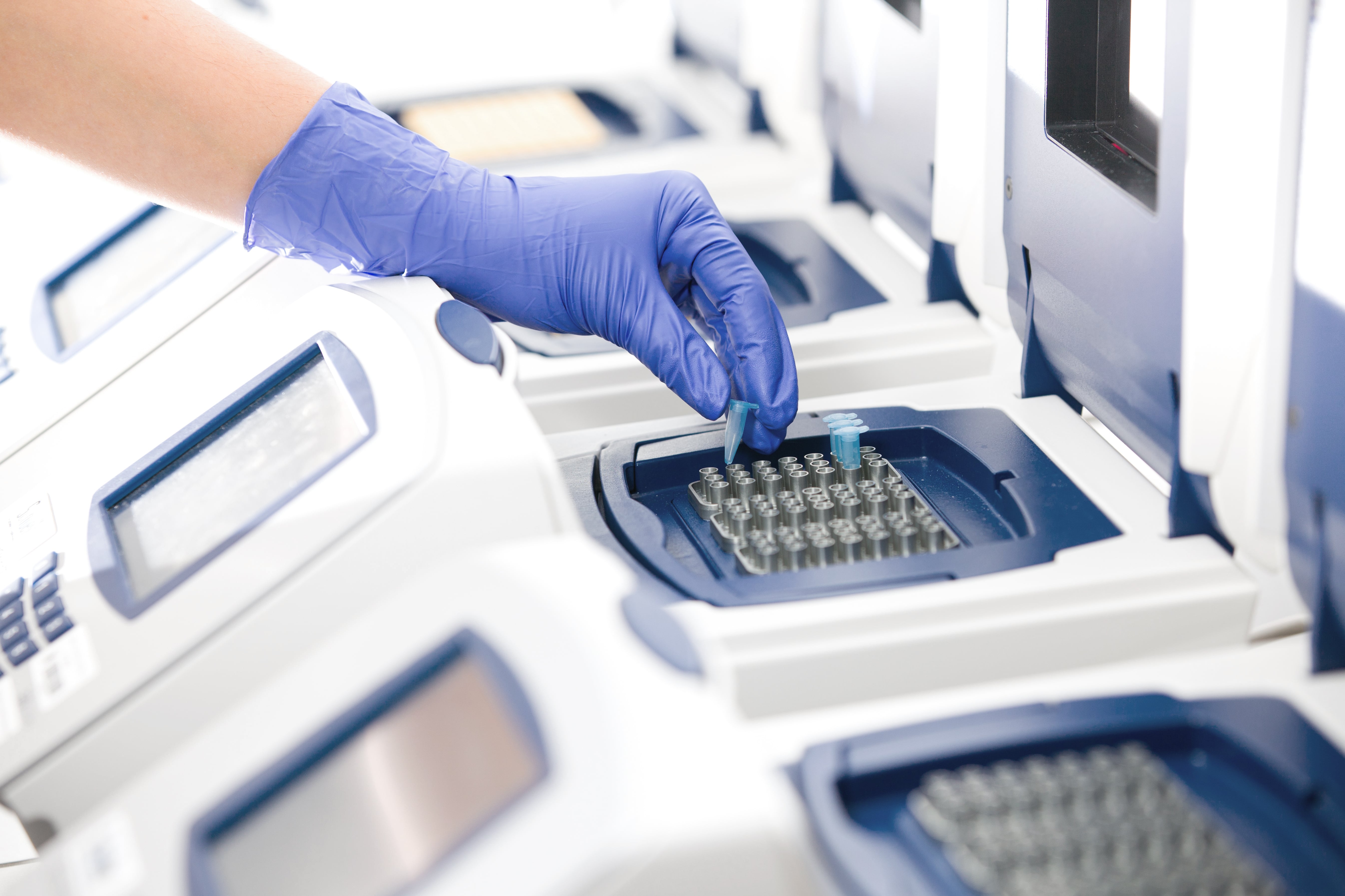 A gloved hand placing a sample tube into a thermal cycler machine used for PCR testing in a laboratory setting.