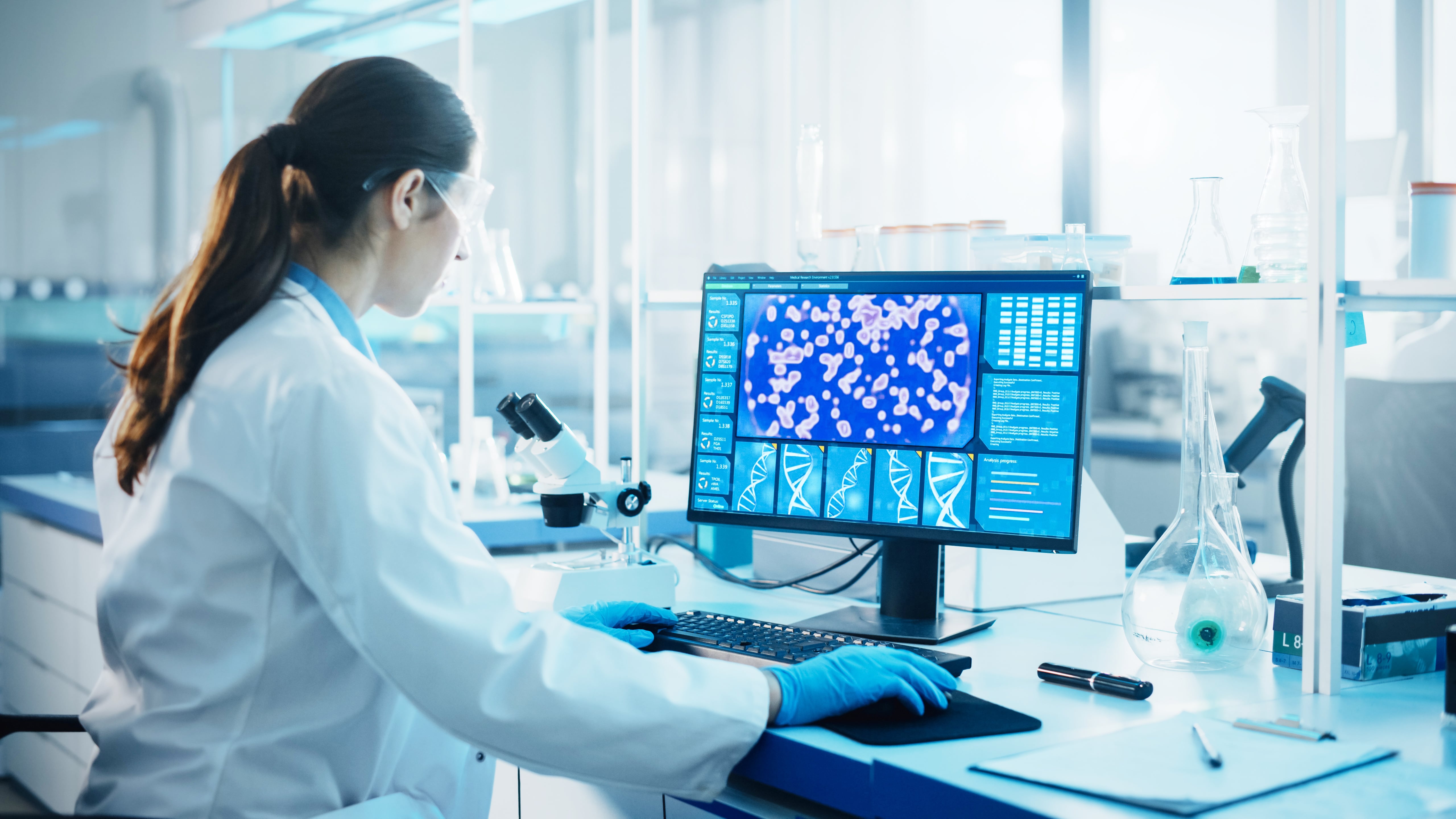 Scientist wearing a lab coat and gloves working at a computer in a laboratory, analyzing microscopic images displayed on the monitor.