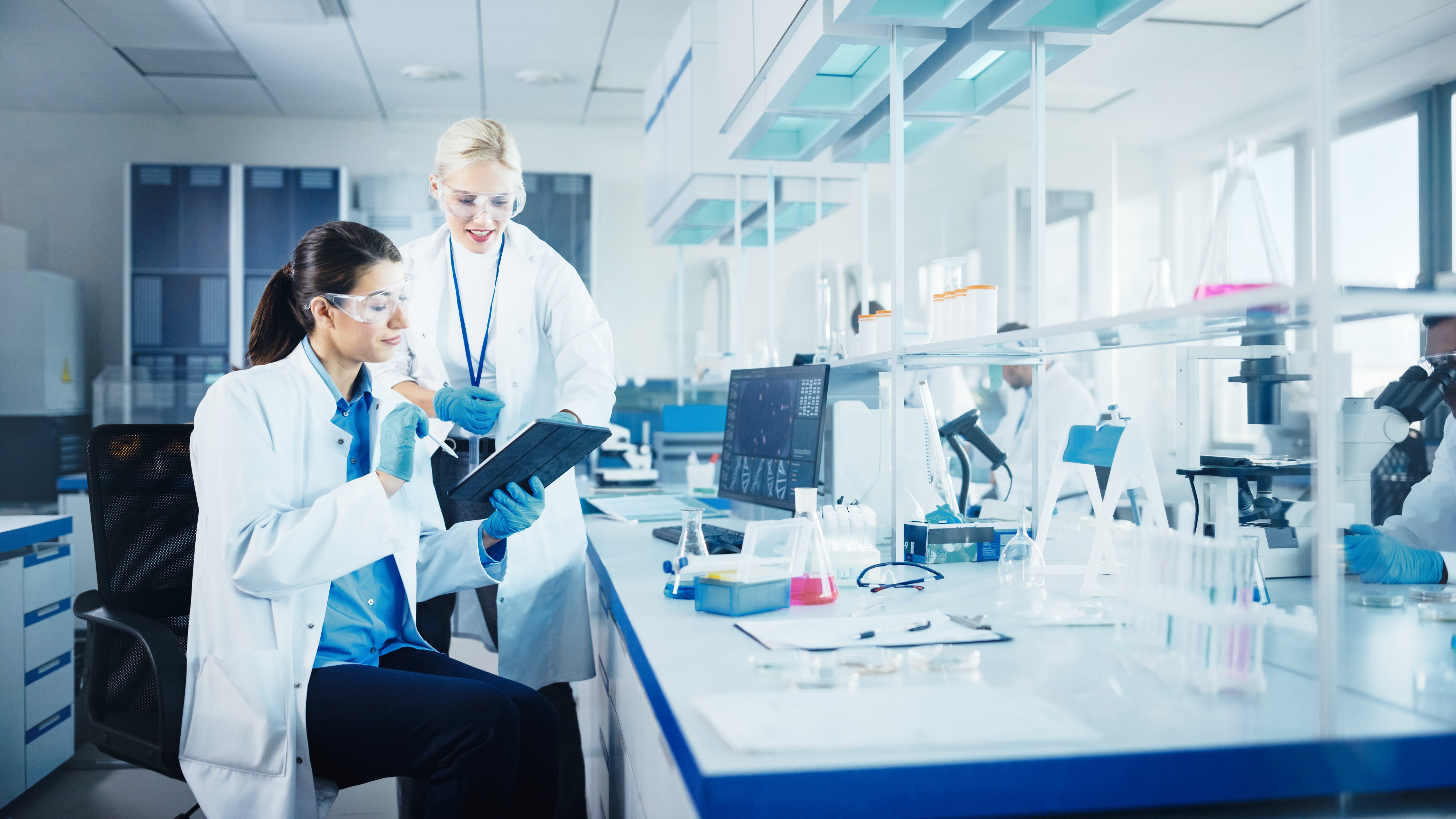 Two scientists in a laboratory wearing lab coats and gloves, working with a tablet and discussing data, surrounded by microscopes, glassware, and computer equipment.