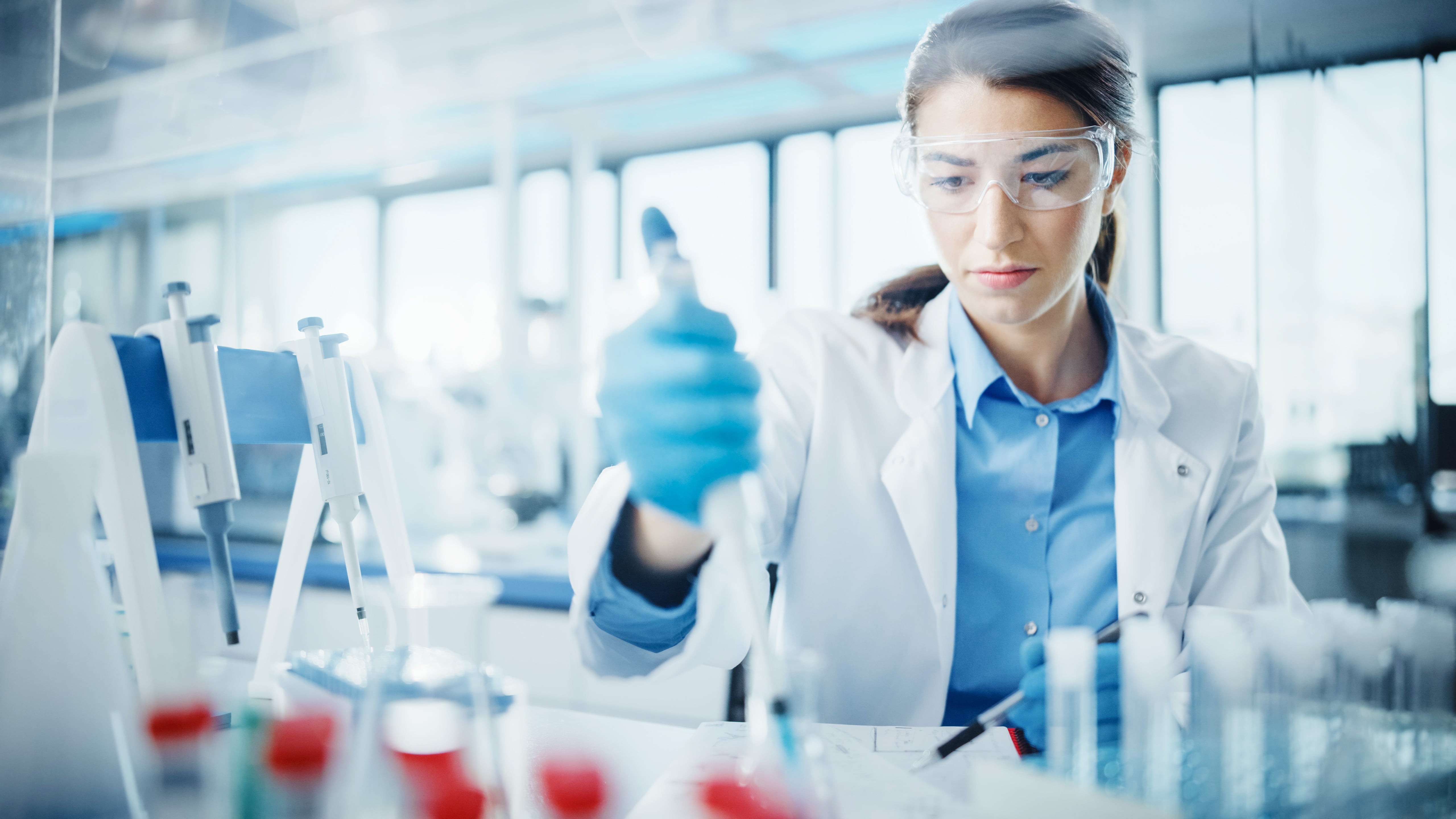 Scientist in a laboratory wearing gloves and a lab coat, holding a pipette and working with test tubes on a bench surrounded by laboratory equipment.