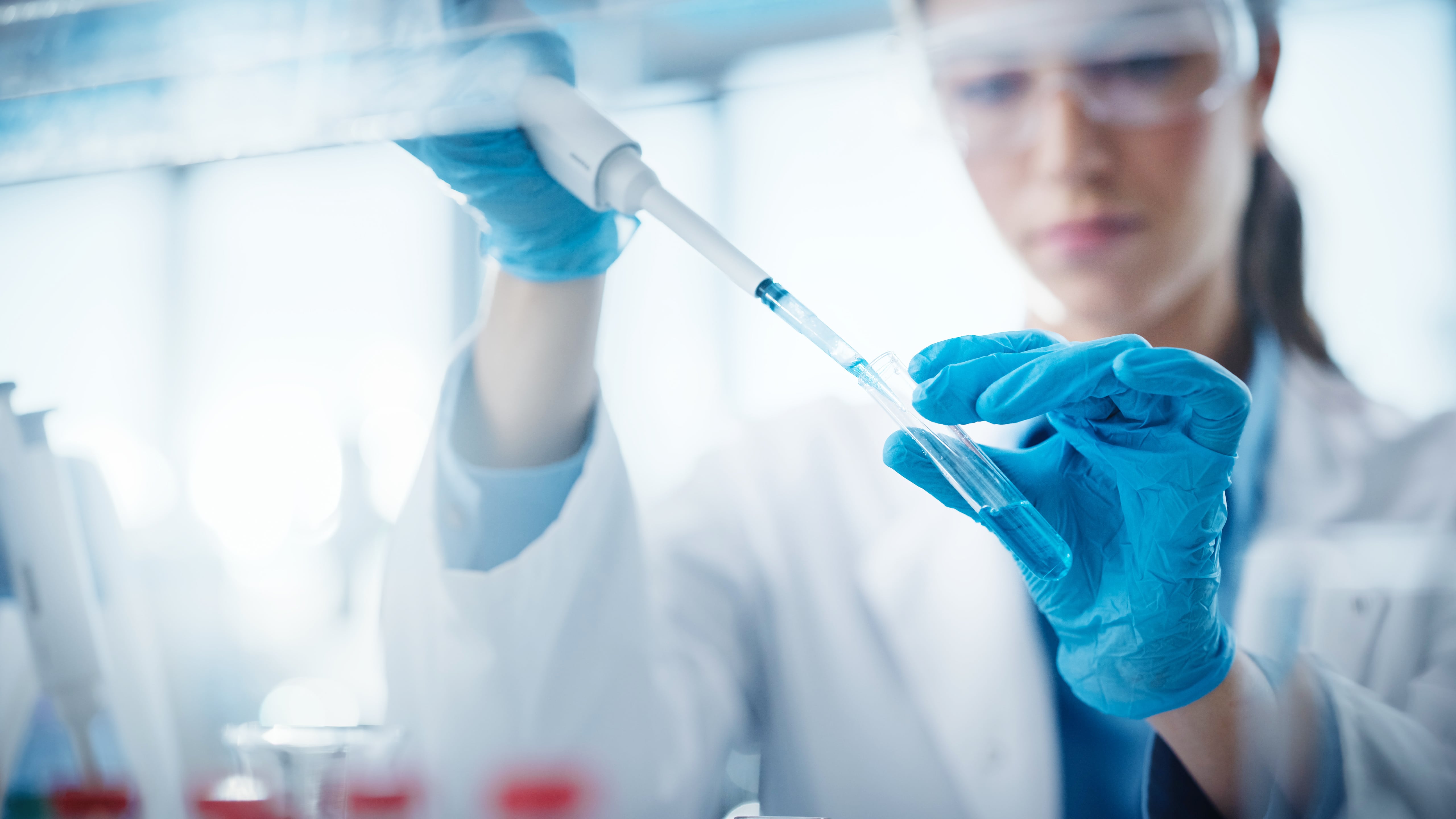 Scientist wearing gloves and a lab coat using a pipette to transfer liquid into a test tube in a laboratory setting.