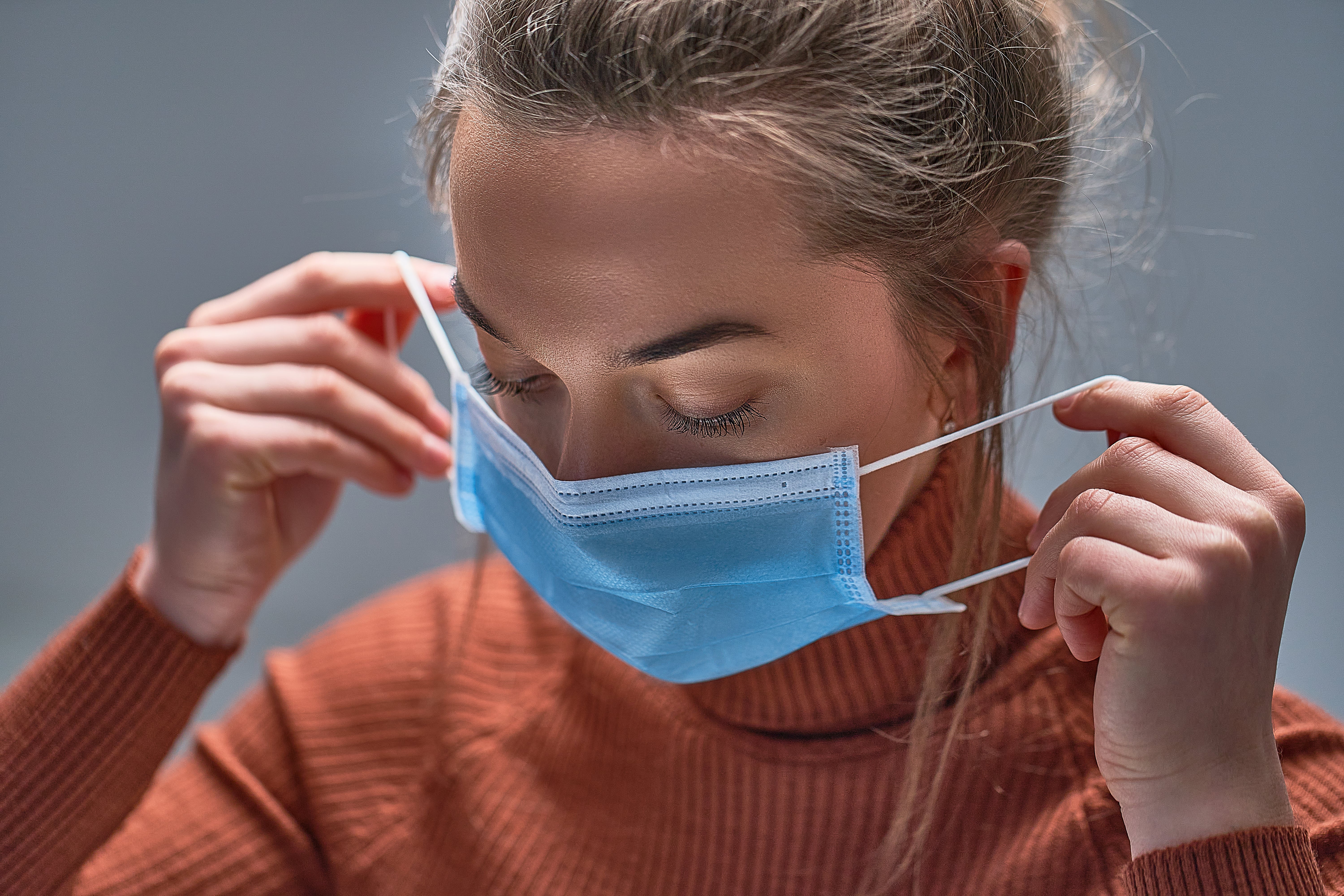 Person wearing an orange sweater putting on a blue surgical face mask using both hands.