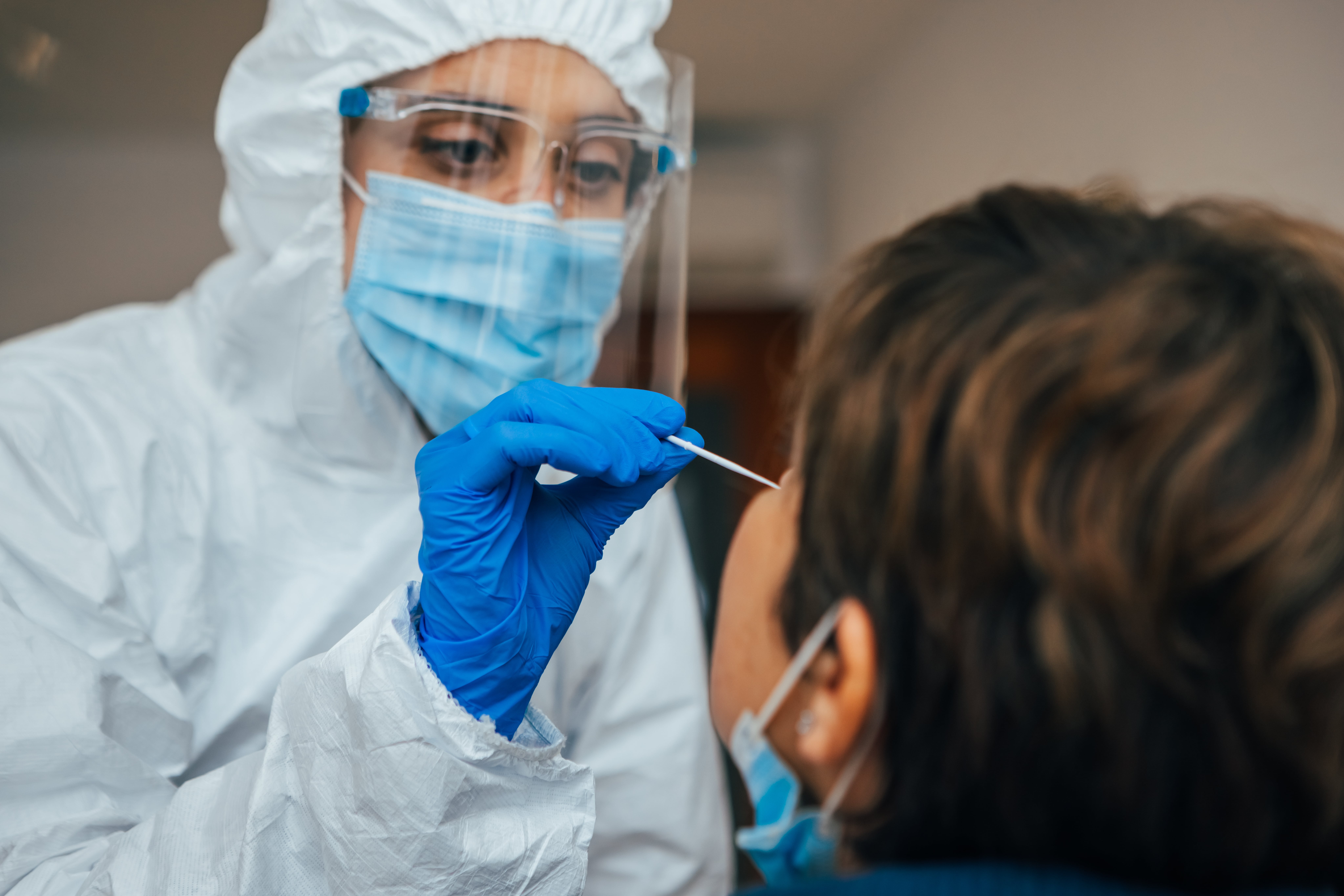 Healthcare professional wearing full protective suit and blue gloves performing a nasal swab test on a person in a clinical setting.