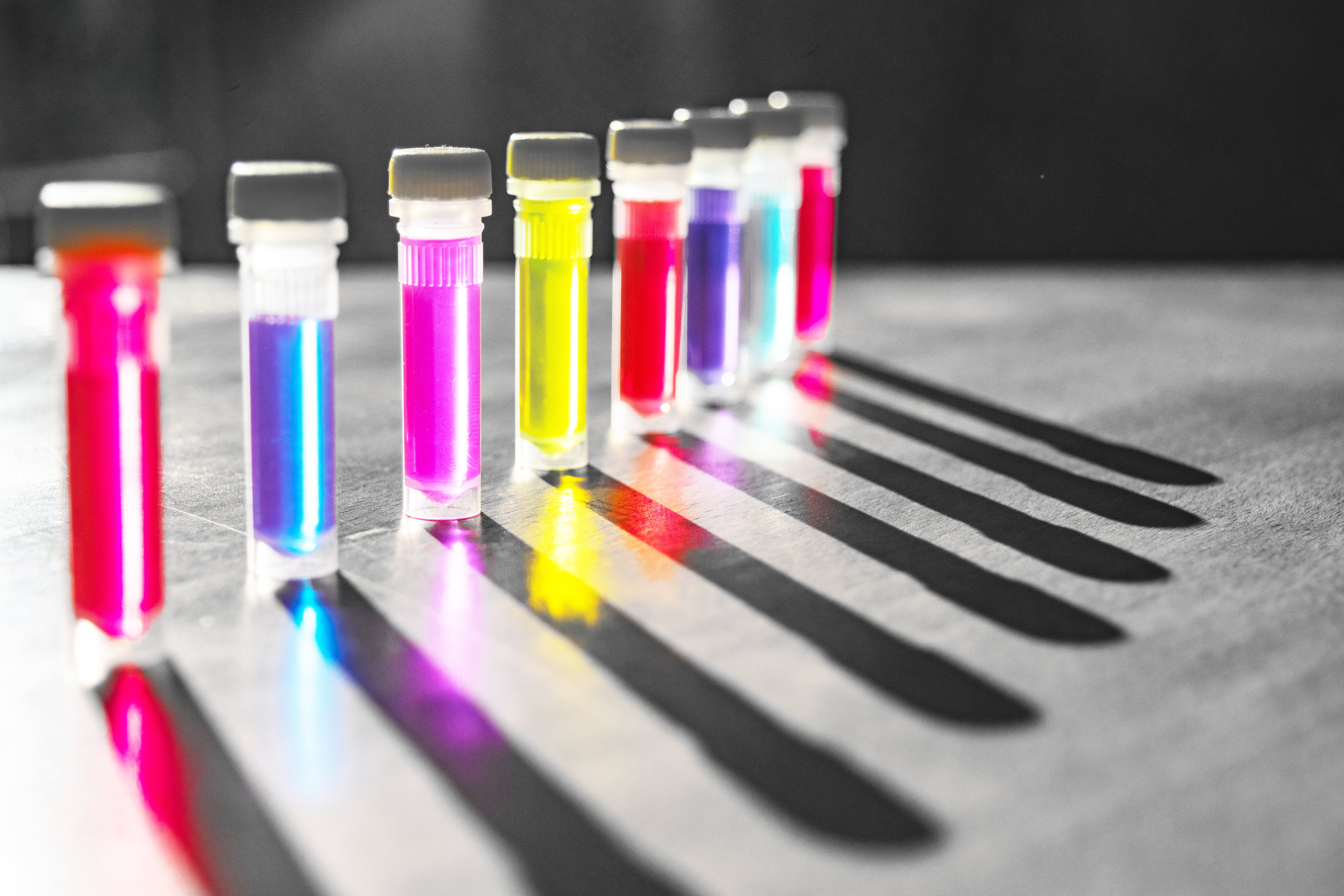 Several small transparent vials filled with brightly colored liquids in shades of pink, blue, yellow, and purple, arranged in a row on a surface with long shadows cast behind them.