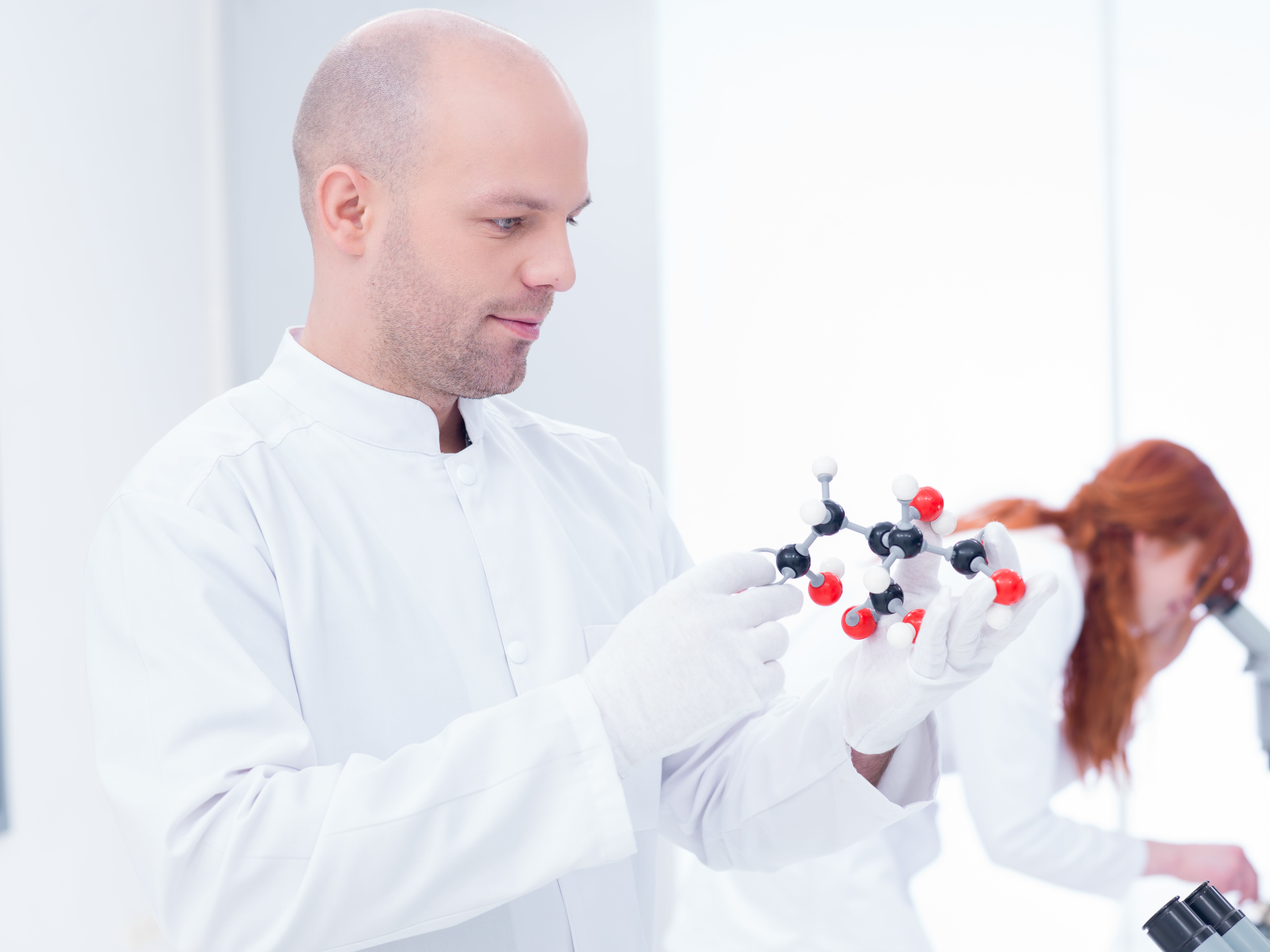 A person in a white lab coat and gloves holding a molecular model in a bright laboratory, with another individual working at a microscope in the background.
