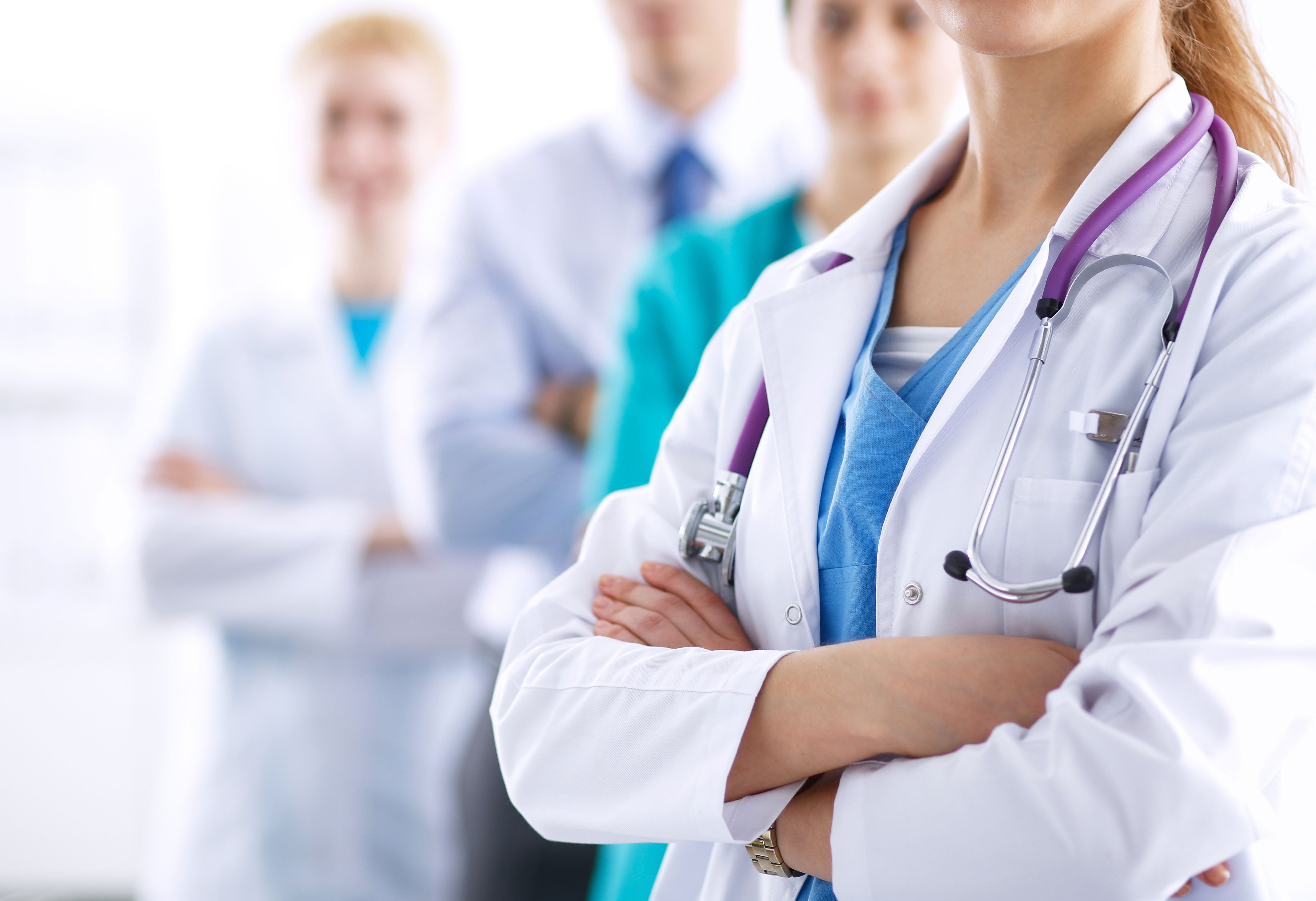 A group of medical professionals in white coats and scrubs, with a close-up focus on a female doctor crossing her arms.