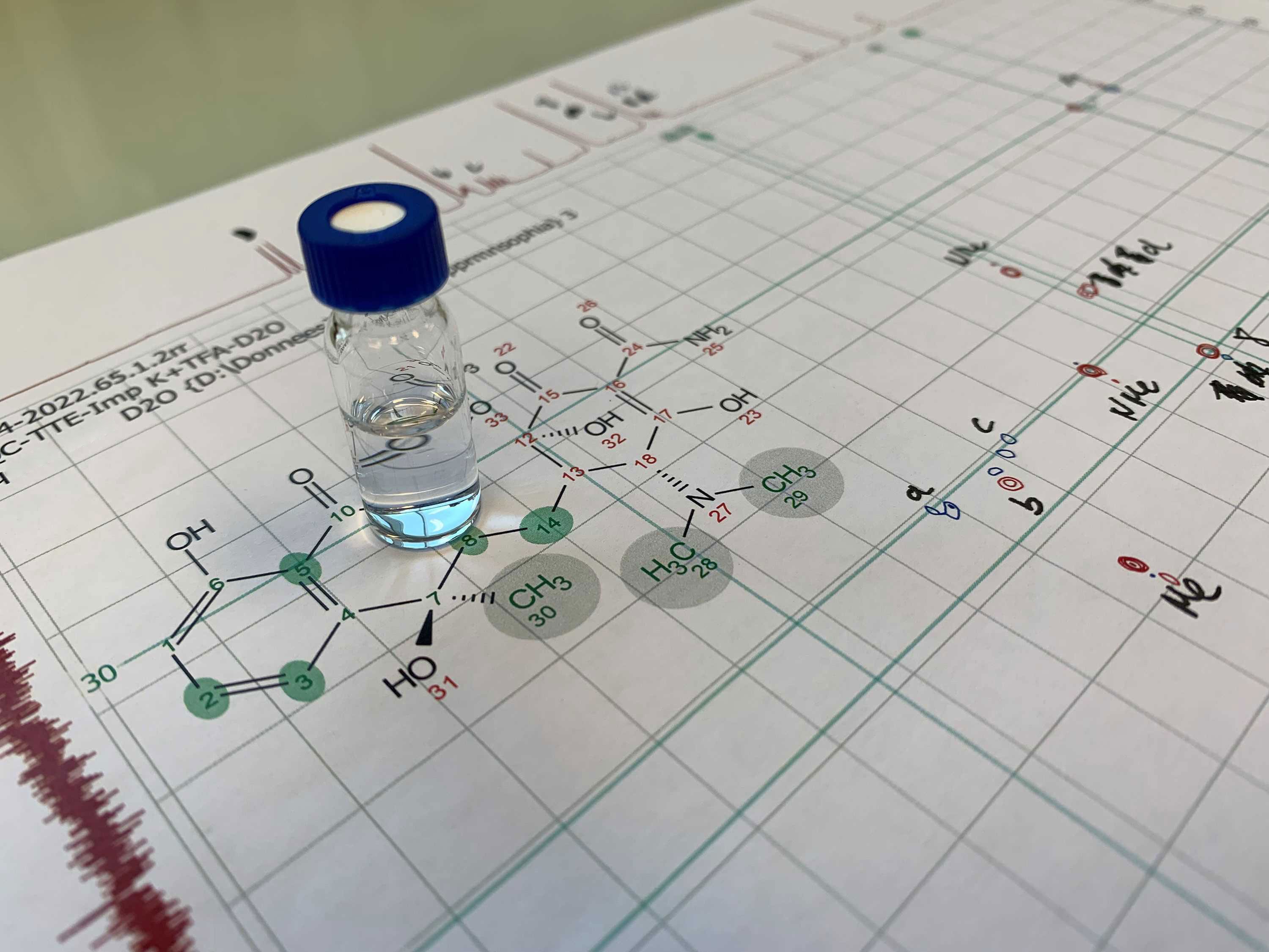 A small vial with blue liquid in it is placed on a spectra paper showing chemical structure of a molecule 