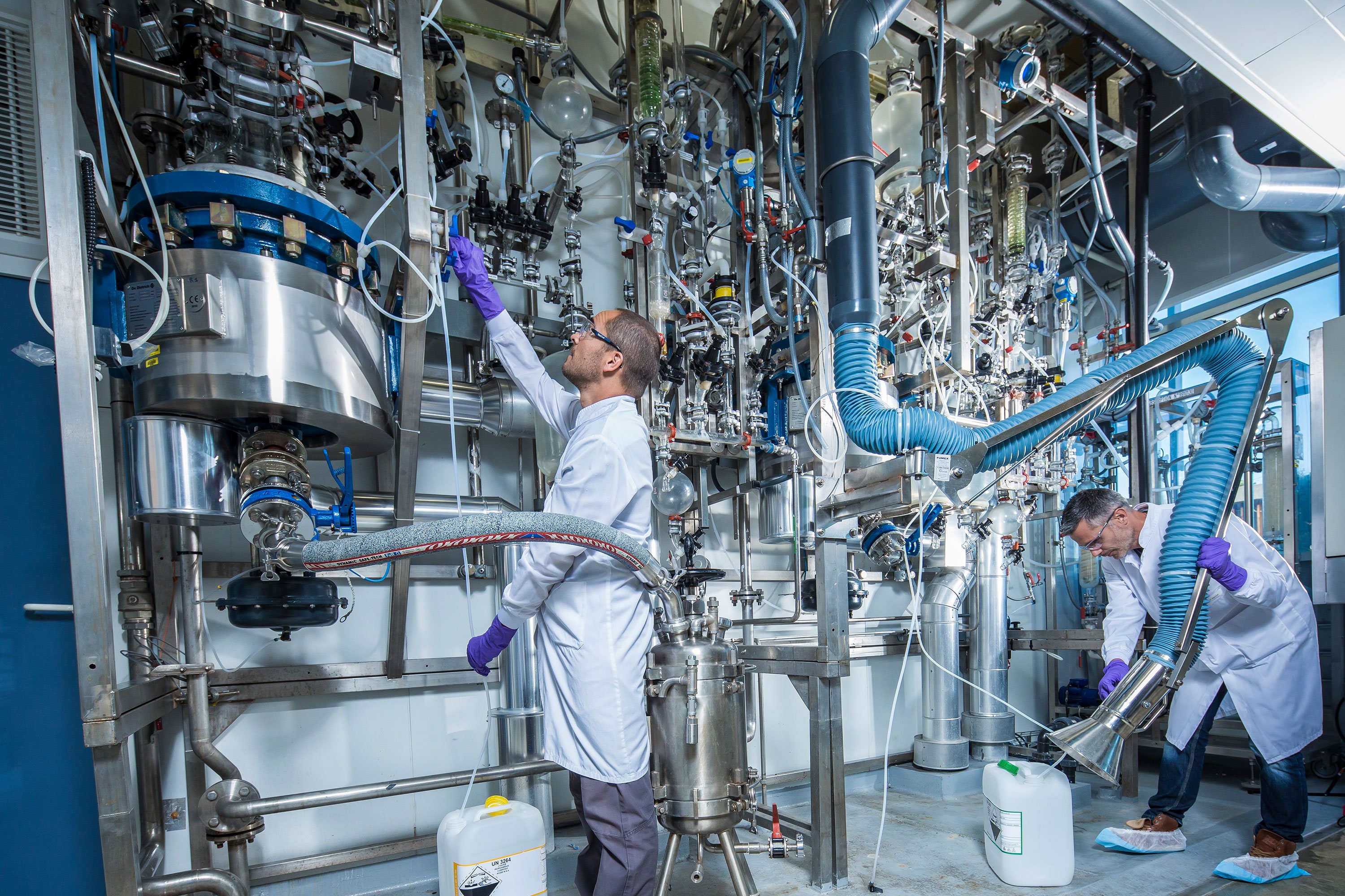 Two lab-coated technicians work in a kilolab—one at a reactor's base, the other adjusting a ventilation tube