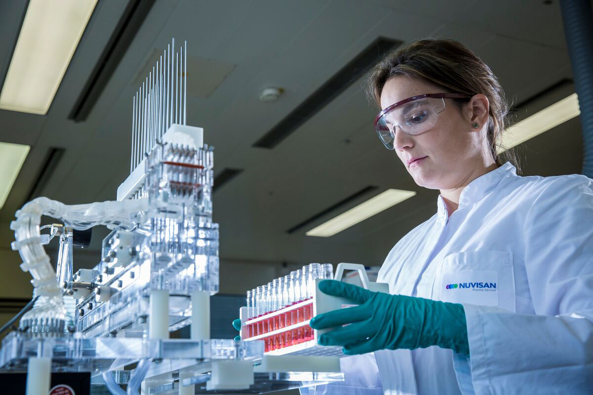 Scientist holds a rack of vials containing red liquid, positioned in front of a multifiller machine in a laboratory setting.