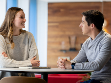 Colleagues engage in conversation across a table in a bright, collaborative office environment.