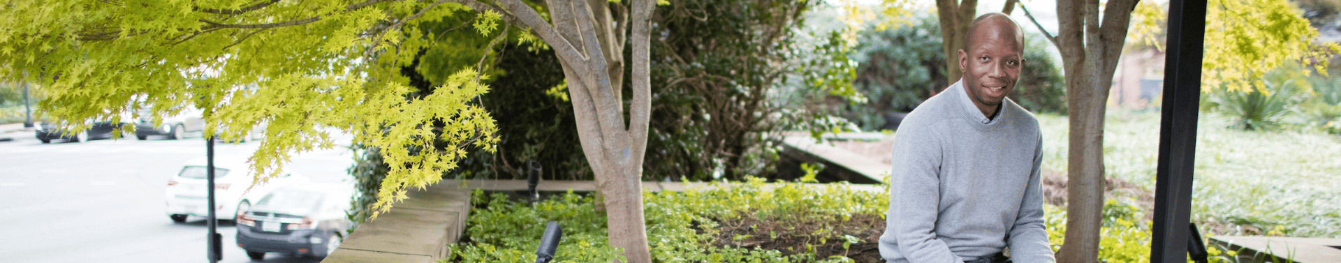 Person seated on a stone planter wall outdoors, holding a phone beneath shaded trees.