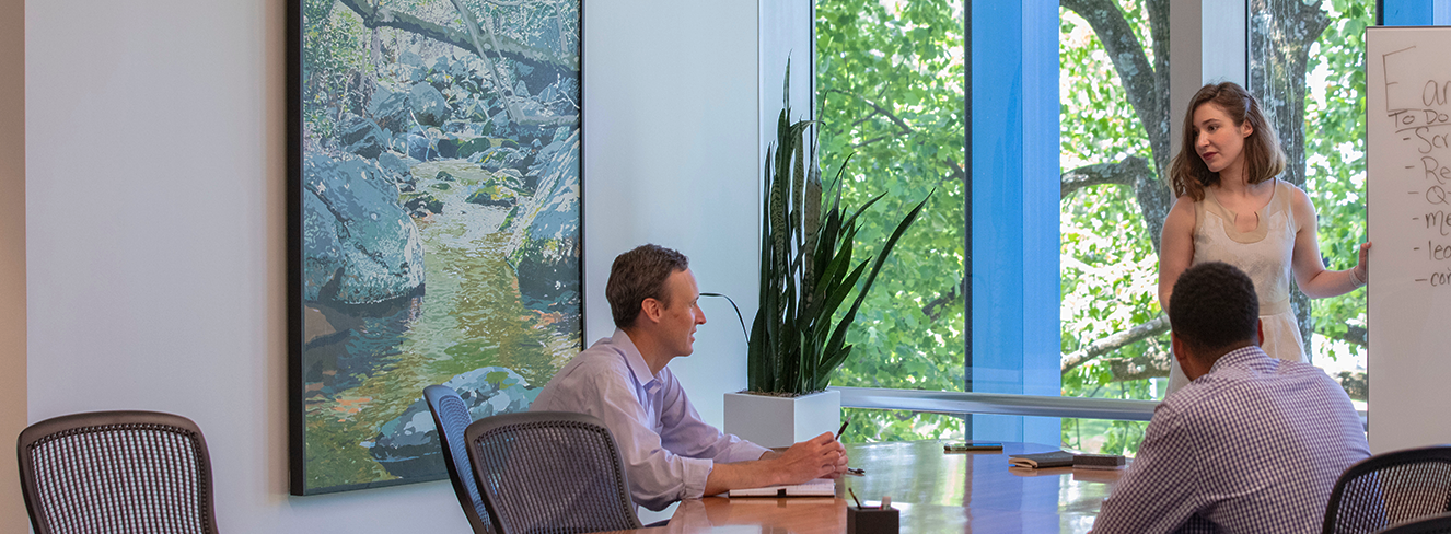 Two males seated at a conference room table with unused chairs around and one female standing before of a glass wall showing trees outside at a whiteboard, while presenting, with a large black framed image of a nature scene against the white walls.