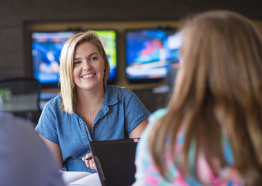 Hiring Process image - Smiling blonde haired white female in a denim blue top facing a female with brown hair with a laptop before her and multiple tv screens in the background