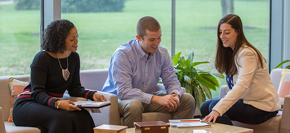 Inclusion, Diversity & Equity image - Two females and one male sitting together in an open lobby area looking at open books on a coffee table before of a glass wall showing grass outside.