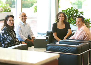 Open Jobs Portal image - Two females and two males smiling into a camera, sitting in an open lobby area on black chairs in front of a glass wall showing the potted plants outside.