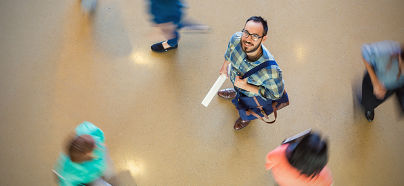 Workforce & Community Capability Building image - Man with close-cropped hair and dark-rimmed glasses in a blue/green plaid shirt with a bag over his shoulder looking up to the ceiling.  People walking around him.
