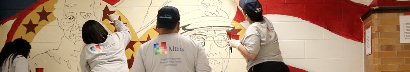 A close-up of four employee volunteers in grey, long-sleeved polos with the Altria logo on the back, painting a mural in red, gold and blue