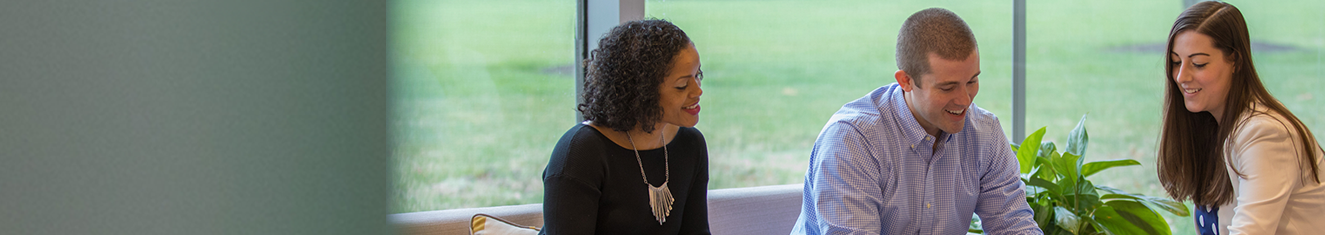 Two females and one male sitting together in an open lobby area looking at open books on a coffee table before of a glass wall showing grass outside.