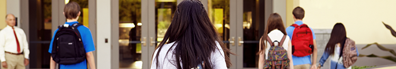 Five students with different colored backpacks in casual dress walking into the front of a school with a male teacher at the left watching their entry