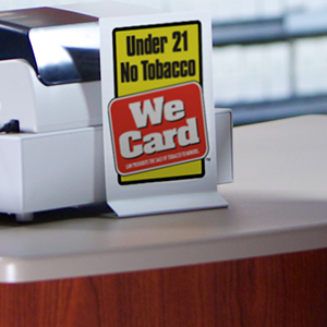 Under 21 No Tobacco We card sign in yellow, red, black and white in a sliver stand in front of a cash register on top of a white counter of a wooden cabinet.