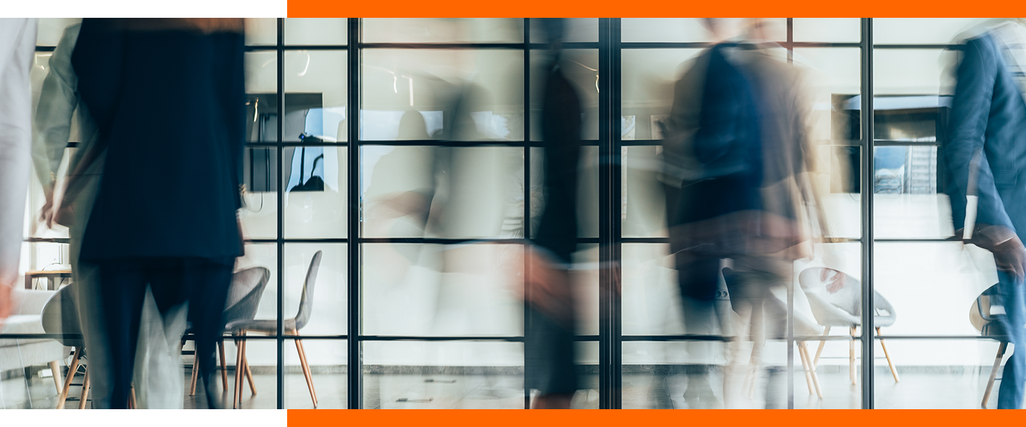Un espace de bureau dans lequel de nombreuses personnes sont en déplacement devant un mur de verre transparent; en arrière-plan, une salle de conférence équipée de deux écrans de télé au mur.