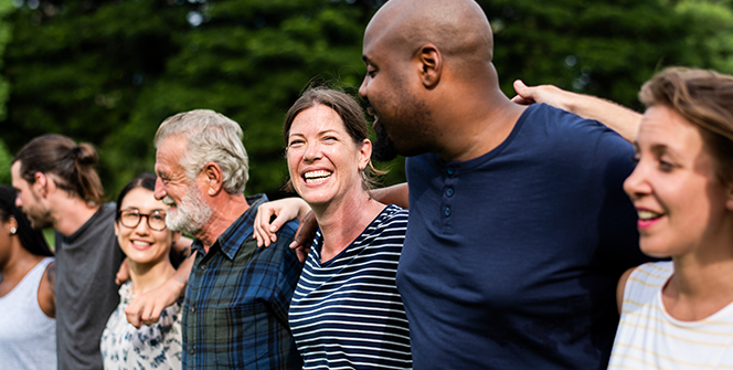 Smiling group of diverse people in a park with arms around shoulders and standing in a long row with a solid bank of dark leafy green trees in the background.