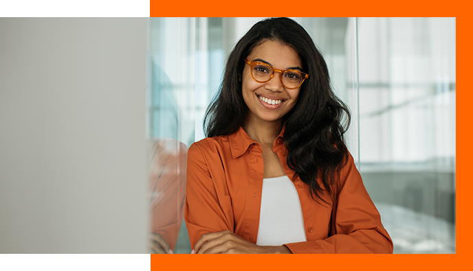 Brown-skinned smiling female with long brown hair wearing tortoise-colored framed glasses and an open burnt orange shirt over a white inner top, leaning against a glass wall.