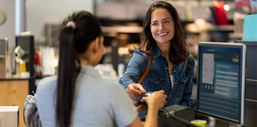 Smiling female with long brown hair standing at a cash register wearing a denim jacket, completing an electronic transaction with a female clerk wearing a light blue short-sleeved shirt with very-long dark hair in a ponytail.
