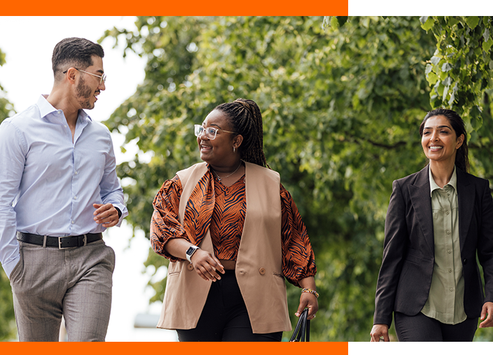 Two female and one male business-people walking outside together, all dressed smartly talking and smiling at each other with a bank of leafy green trees in the background.
