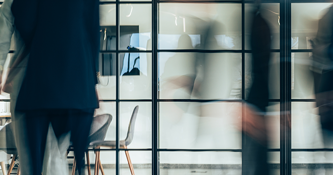Un espace de bureau dans lequel de nombreuses personnes sont en déplacement devant un mur de verre transparent; en arrière-plan, une salle de conférence équipée de deux écrans de télé au mur.