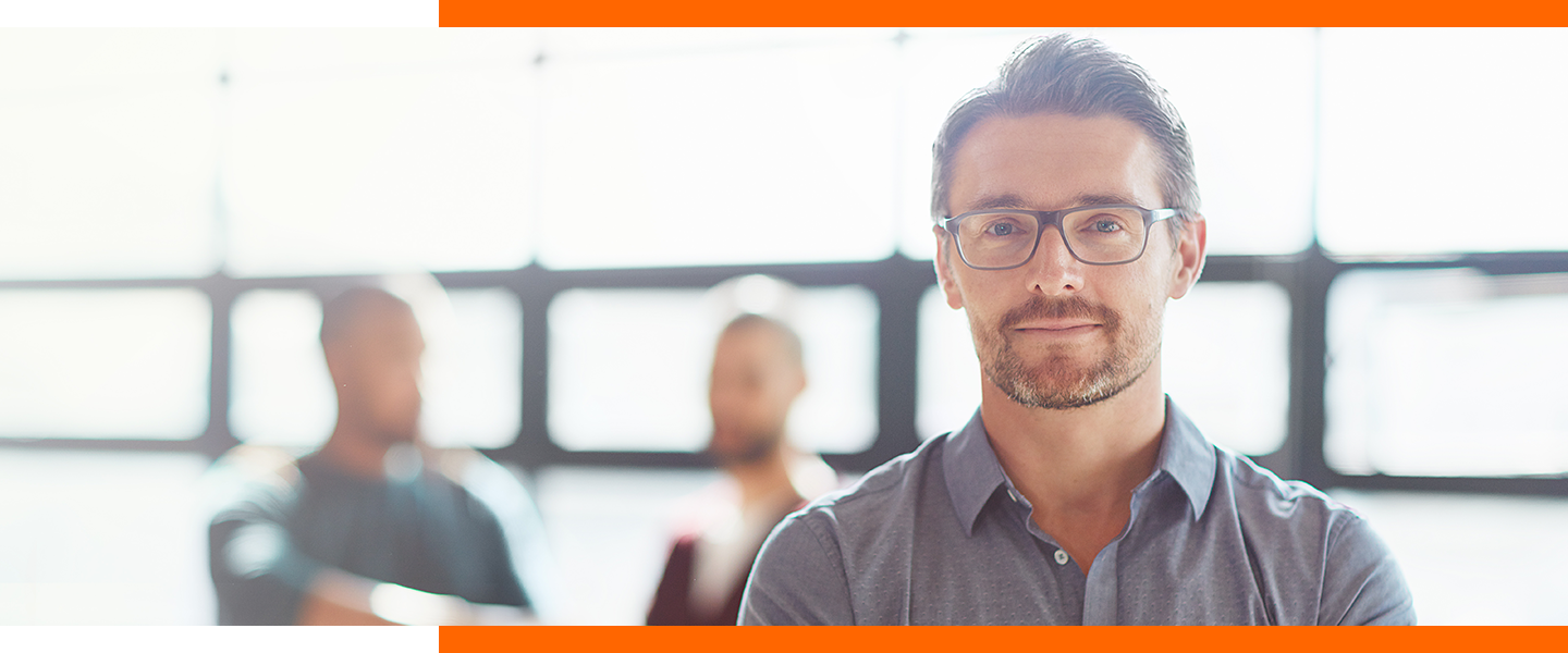 Un homme blanc barbu aux cheveux bruns courts, portant une chemise boutonnée gris ardoise et des lunettes à monture foncée, debout dans un bureau avec ses collègues en arrière-plan, devant un mur de fenêtres où le soleil pénètre à flots.