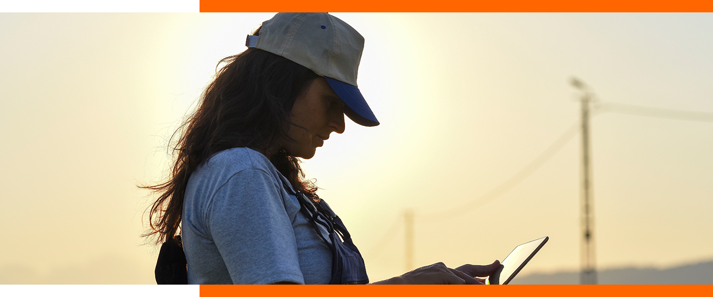 Shadowed side-view of a female with long wavy dark hair, wearing a white cap with a blue bill, standing in a field and focused on an tablet with a line of electrical poles in the background.